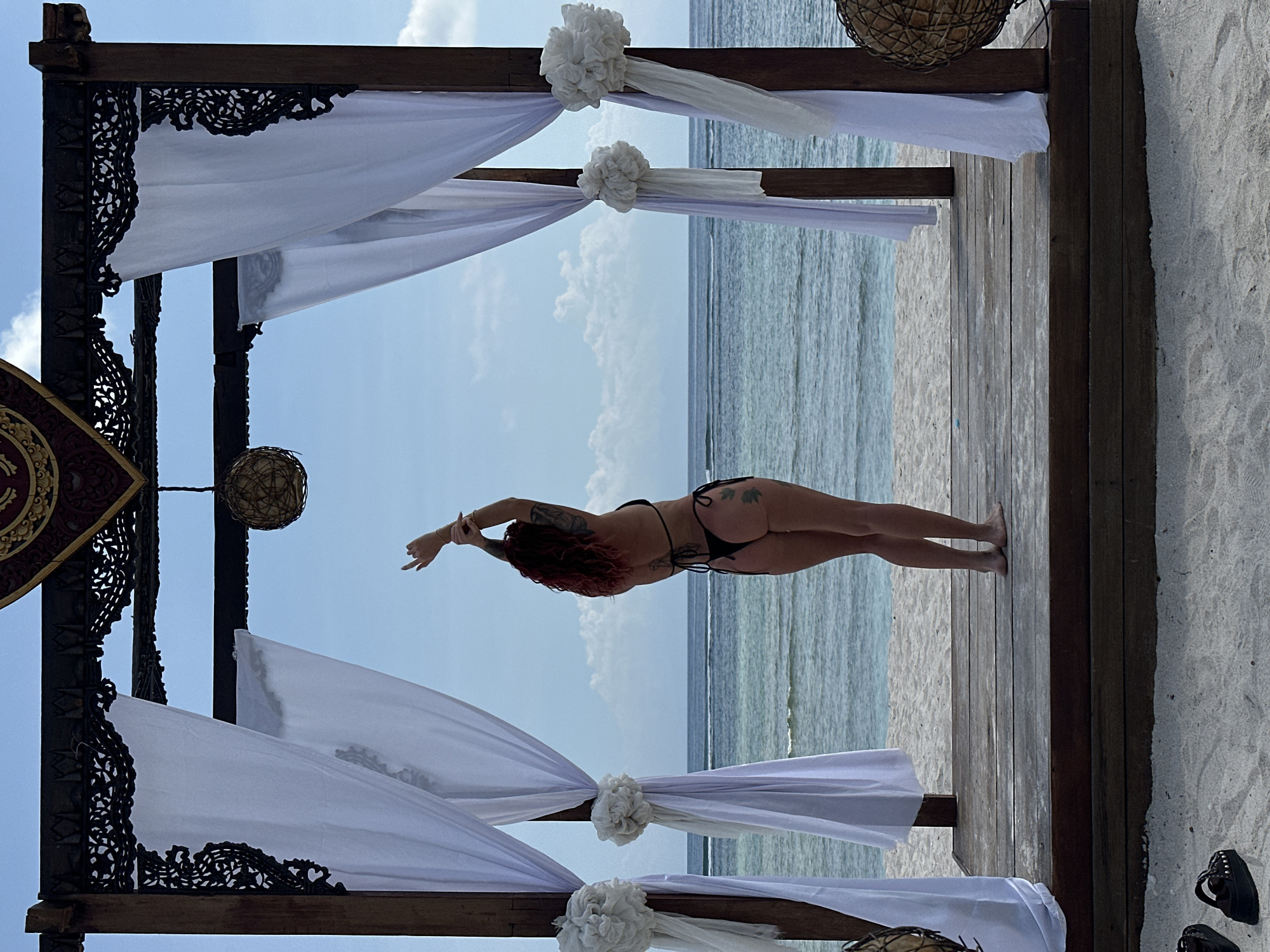 The photograph depicts a woman in a black bikini standing on a wooden platform. Her arms are extended. She has a tattoo on her back and upper arms. The wooden platform leads out to a beach and the ocean. Above and around the woman is a wooden structure draped with white fabric and adorned with floral decorations. The structure features intricate carvings. A decorative object hangs from the structure. The location appears to be on a beach, possibly in Indonesia.