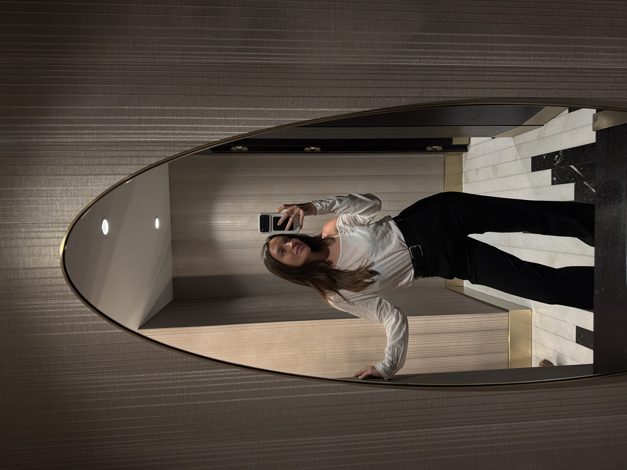 The image captures a full-body mirror selfie of a young woman within an interior space located in Budva, Montenegro. The woman, with long, dark hair, stands centered in the reflection, smiling gently as she looks towards her smartphone. She is dressed in a light-colored, off-the-shoulder long-sleeved top and black high-waisted trousers secured with a black belt featuring a silver buckle. Her right hand holds a white smartphone with a prominent white case, positioned to capture the reflection. Her left hand rests on the thin, gold-colored frame of the large, irregularly shaped mirror.
The surrounding environment, visible both directly and through the mirror's reflection, features walls covered in a textured wallpaper with subtle vertical stripes in a muted taupe or beige tone. The floor, primarily visible in the reflection, is composed of rectangular tiles in a alternating pattern of white and black, some exhibiting a marble-like veining. Gold-colored trim or baseboards are present at the juncture of the walls and floor in the reflected space. Overhead, two recessed circular lights are visible on the ceiling in the reflection. A dark-colored door frame or architectural partition with dark wood elements is also partially reflected in the background, contributing to the impression of a modern and well-appointed indoor setting.