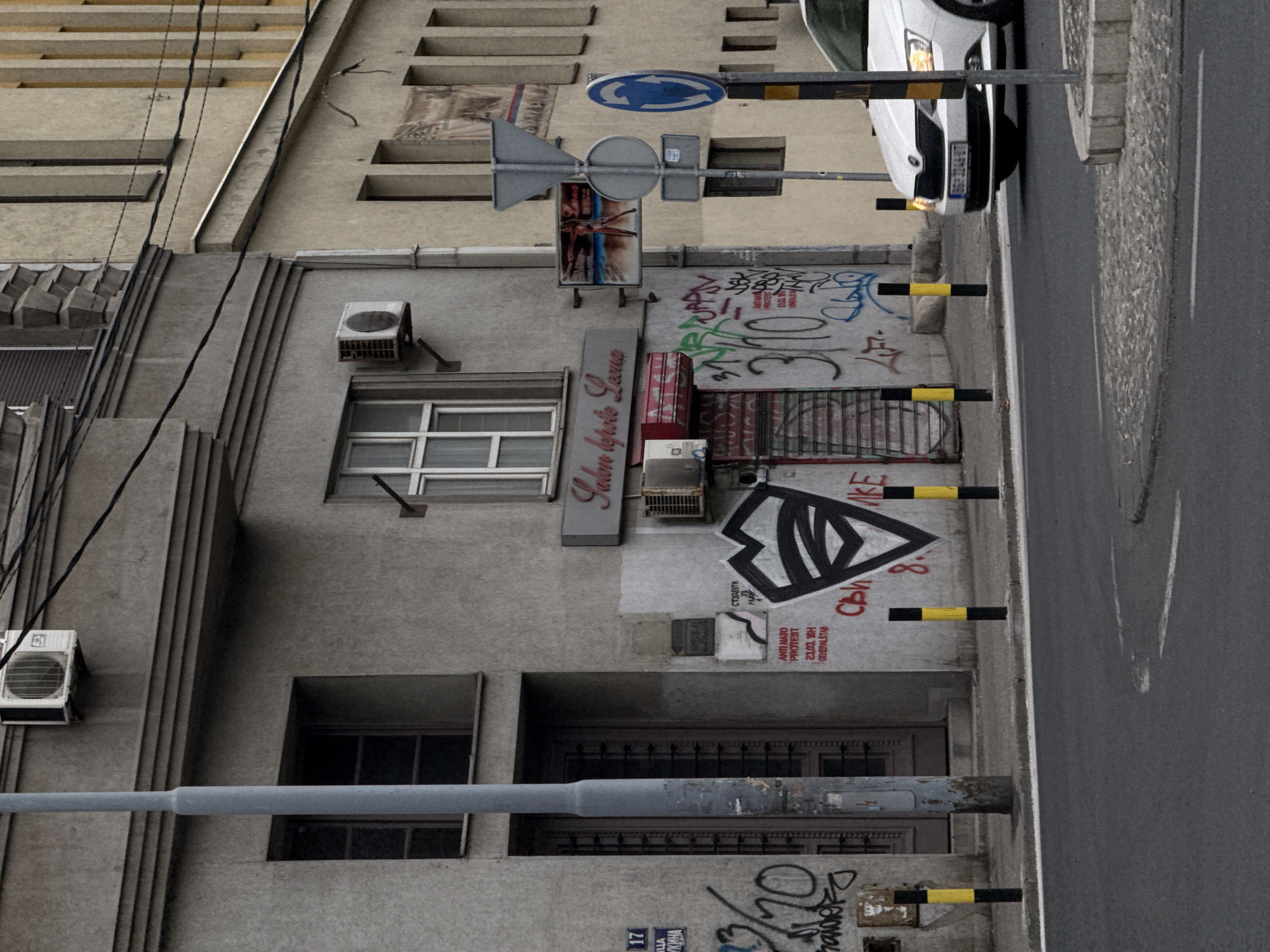 This rotated image depicts a gritty, urban street scene in Belgrade, Serbia, featuring a building facade, a street, and a parked car under an overcast sky during daytime. The primary subject is a multi-story concrete building with a commercial establishment on the ground floor.
In the foreground, a gray utility pole extends horizontally across the frame. Above it, a building shows multiple windows, some open, and an air conditioning unit. The street level features a sidewalk with black and yellow bollards, separating it from an asphalt road. A white car, possibly a Skoda, is parked facing right on the roadside, with its front end and partially visible "BG" (Belgrade) license plate number.
The building's facade is heavily marked with graffiti, including large, abstract designs and text. A sign for "Salon Lepote Leona" (Beauty Salon Leona) is prominently displayed. Other smaller signs and posters are visible, one of which explicitly states "ANTI NATO PROTEST 23.03.16H GENERALSTAB" (Anti NATO Protest, March 23rd, 4 PM, General Staff building), providing a significant historical and political context to the location. Another piece of graffiti reads "17 ЦА КИНА" (17 TSA KINA), possibly indicating a street name or address. A blue circular road sign with a white right-turn arrow, and another gray rectangular sign, are attached to a pole above the street. Overhead, numerous electrical wires crisscross the frame. The overall atmosphere is one of a busy, lived-in urban environment with visible signs of local culture and history.