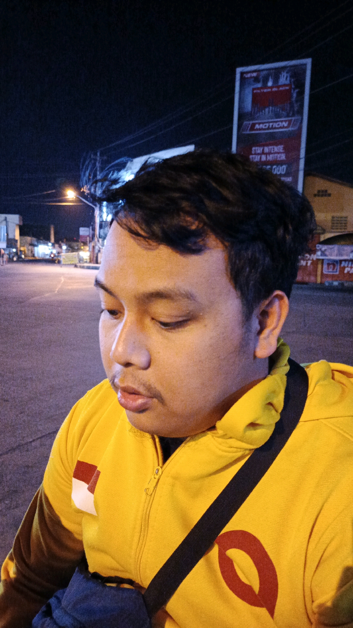 A close-up, medium shot, taken at night, shows a young man from the chest up, looking down and slightly to his right. He is wearing a bright yellow hooded jacket with a black strap across his chest and a dark blue bag slung across his shoulder. A red and white emblem is visible on the left side of his jacket. The background shows a street scene in Surakarta, Indonesia, with streetlights, buildings, and a billboard in the distance. The billboard has the word "MOTION" prominently displayed, along with text that appears to be in Indonesian. The man's face is illuminated by artificial light.