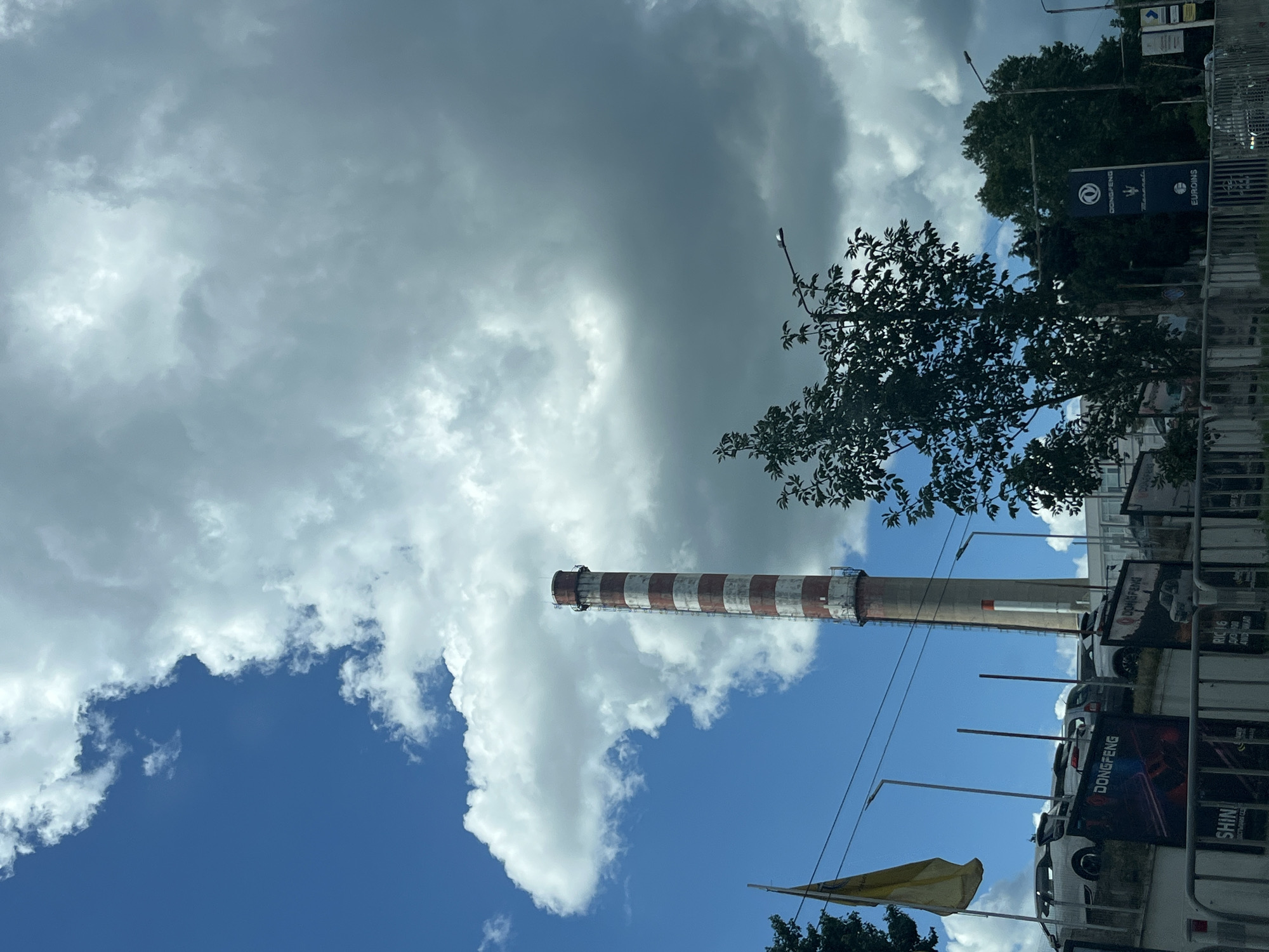 The image depicts a street view in Varna, Bulgaria, with a prominent industrial chimney in the background. The sky is partly cloudy, with a mix of bright blue sky and heavy gray clouds. The chimney is tall and cylindrical, painted with alternating red and white horizontal stripes. In the foreground, there is a row of cars parked behind a fence, possibly a car dealership, with signs indicating "DONG FENG" and other brands. Trees are present on the right side of the image, obscuring some of the buildings behind them. A lamppost is visible above the trees. A yellow flag is flying on the left side of the image, attached to a pole. The overall scene suggests an urban or industrial area on a somewhat overcast day.