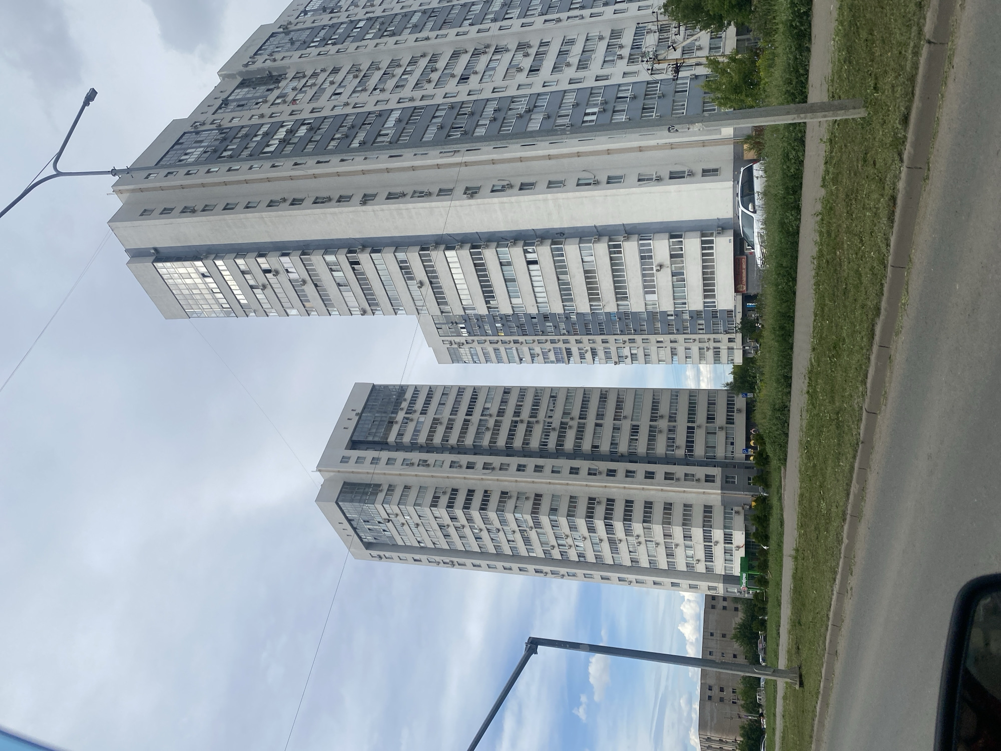 The image depicts a street view in Samara, Russia, focusing on two large, modern apartment buildings. The building on the left is taller and thinner, with a light grey facade and numerous windows. The building on the right is wider and also grey and white, featuring a prominent blue stripe running vertically down its side. Both buildings have many stories and balconies, with visible air conditioning units attached to many windows. In the foreground, a paved road with a grassy median separates the viewer from the buildings. Several cars are visible parked in front of the apartment buildings, and a green grocery store sign is present on the ground floor of the wider building. The sky is overcast with some patches of blue and white clouds. A lamppost with streetlights extends into the upper right corner of the frame, and power lines are visible crossing the sky. The overall impression is of a contemporary urban residential area under a cloudy sky.