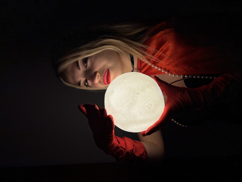 The image displays a close-up, horizontally oriented portrait of a person, likely a woman, in a dark, low-light setting. The main subject is positioned diagonally, with her head and upper torso visible in the upper portion of the frame, looking downwards and to the right.
She has long, light blonde hair that cascades around her shoulders. Her face is softly illuminated by a glowing object she holds. She wears bright red lipstick and her eyes are looking towards the glowing sphere.
She is dressed in a red garment, possibly a top or a dress, which is visible on her right shoulder and arm. Around her neck, she wears a black choker, and a string of white pearls is draped across her chest and shoulders, partially obscured by her hair and the red fabric. Her hands are adorned with long, elegant red gloves that extend past her wrists.
The central focus of the image is a spherical, illuminated lamp designed to resemble the moon, complete with textured craters and variations in brightness, giving it an authentic lunar appearance. This "moon lamp" is held delicately in her gloved hands, positioned centrally in the image. The lamp emits a soft, warm, whitish-yellow light, which acts as the primary light source for the scene.
The light from the moon lamp casts a glow on her hands, the red gloves, the pearl necklace, and subtly illuminates her face, highlighting her features and the red lipstick. The background is almost entirely black, creating a strong contrast and emphasizing the subject and the glowing object. The overall mood is mysterious, enchanting, and dramatic, with the dark setting drawing attention to the illuminated elements. There is no visible text in the image.