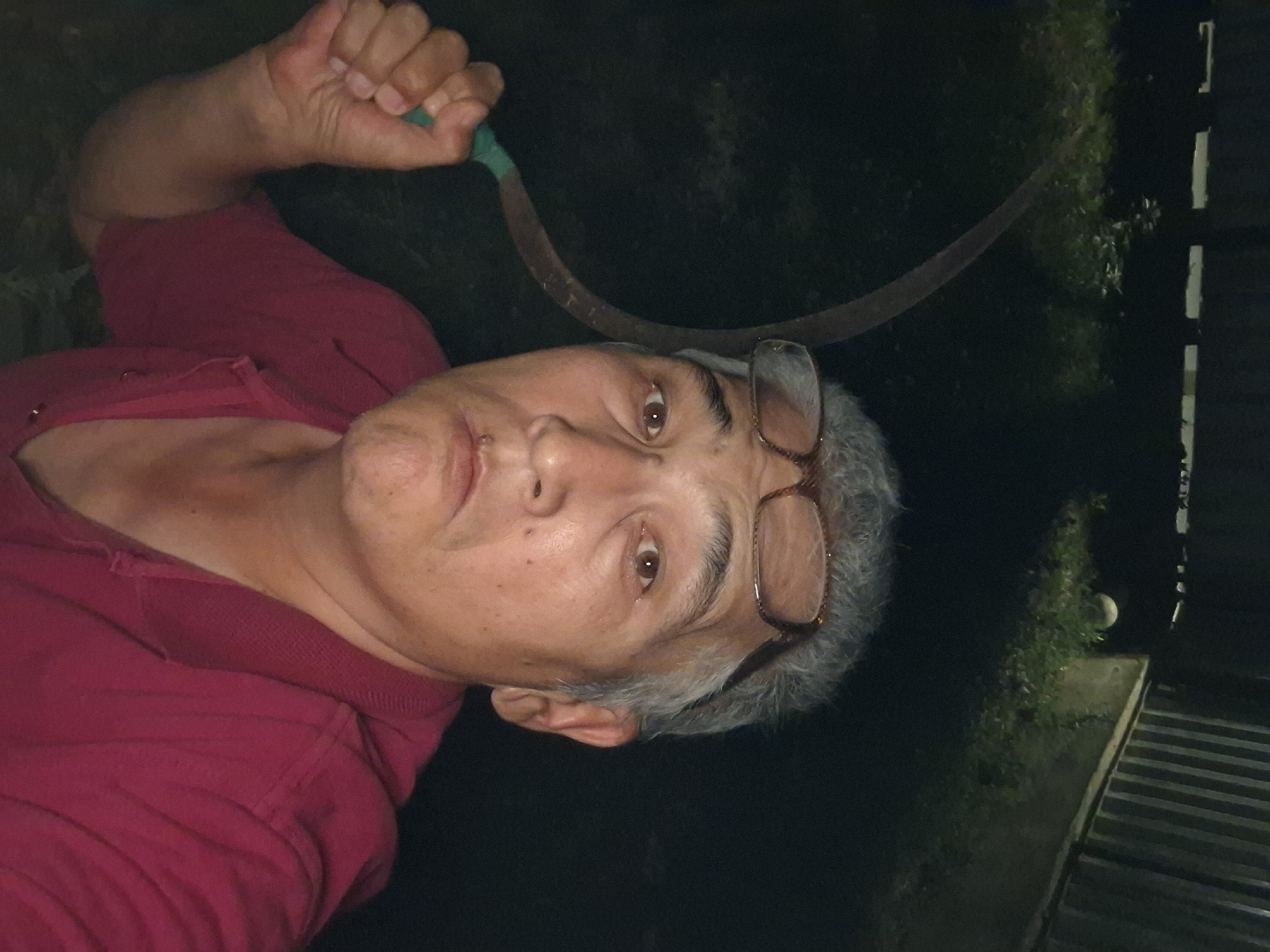 A man, appearing to be of Asian descent, is captured in a low-light, outdoor selfie. He has short, graying hair and is wearing eyeglasses perched on top of his head. The man is holding a sickle, with its curved metal blade visible and a green handle in his right hand. He is wearing a dark red collared shirt. The background is dark, suggesting it is nighttime, with some indistinct vegetation and a fence-like structure visible. There is a faint glow of light from the right side of the image. The setting appears to be a yard or garden area.