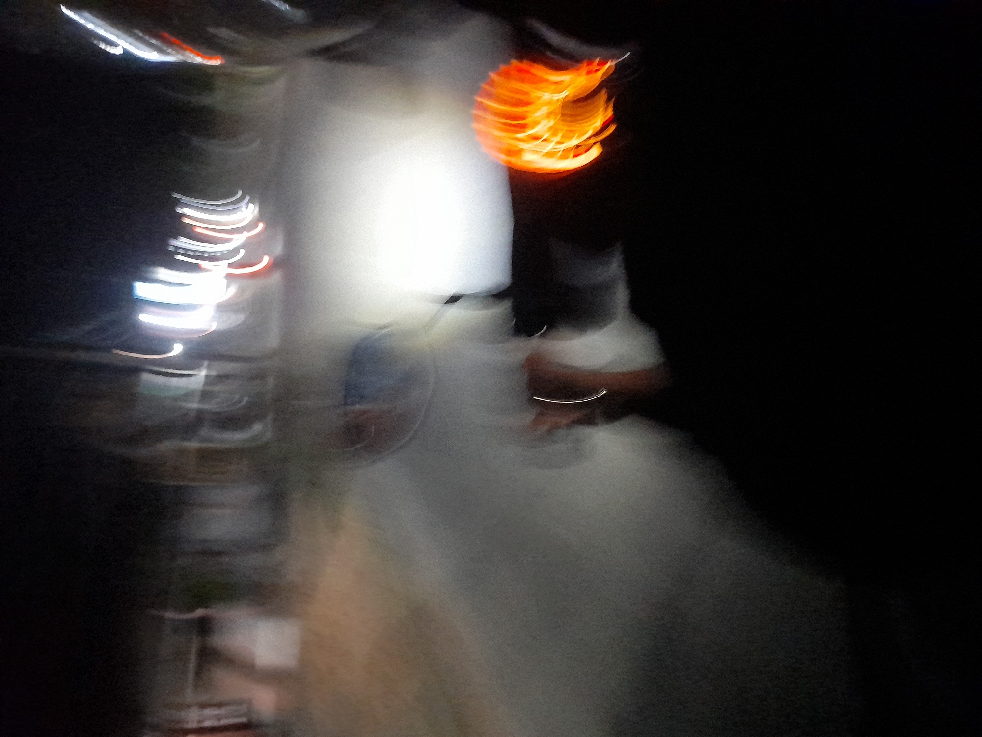The image depicts a blurred nighttime scene, likely captured from the perspective of someone riding a vehicle. Streaks of light from streetlights and possibly other vehicles create a sense of motion. In the foreground, an orange, circular light source, possibly a taillight or a reflective object, is prominently visible. The background shows indistinct shapes of buildings or structures, also blurred due to movement and low light conditions. The overall impression is one of a fast-moving vehicle on a road at night in a populated area. Without further information or sharper detail, specific identification of locations within Jetaksari, Indonesia is not possible from this image alone.