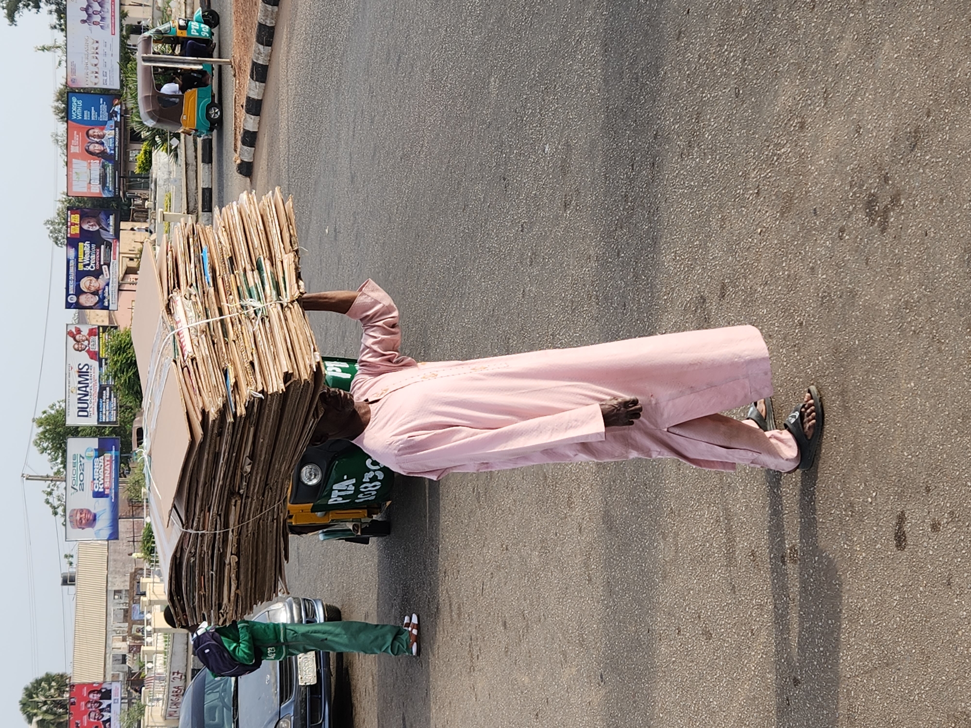 The image depicts a man carrying a stack of cardboard on his head in Jos, Nigeria. The man is wearing a long pink robe and sandals. He is walking on a paved street. Behind him, there are cars and billboards, including one for Dunamis International Gospel Centre. A three-wheeled vehicle is parked nearby. Another man in green trousers is standing to the left. Billboards visible in the background promote Chris Kwaja for Senate and feature advertisements for religious events. The license plate on a vehicle is visible.
