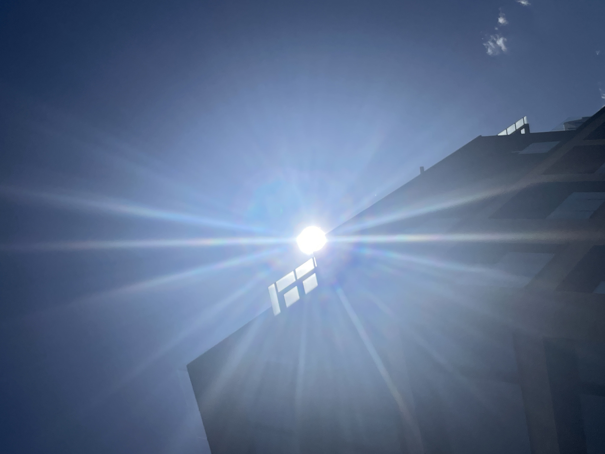 A low-angle, daytime shot shows a structure against a blue sky with the sun positioned behind it. A bright, intense glare emanates from the sun, creating a lens flare effect. The structure appears to be a modern building with what seems to be a partially visible, grid-like pattern of windows or architectural elements. The building's material is dark, causing it to appear silhouetted. A few wisps of clouds are visible in the upper right corner of the frame.