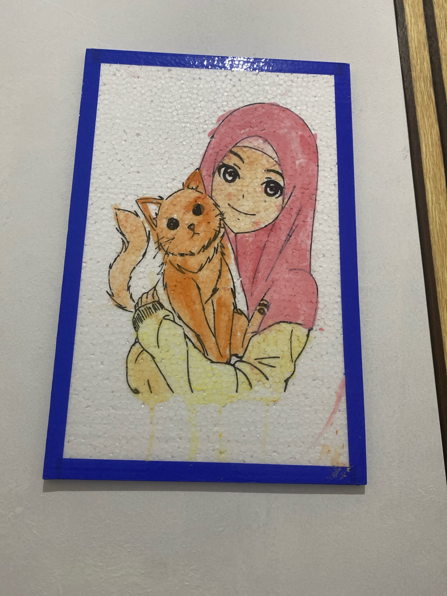 This image displays a piece of artwork featuring a young person wearing a pink hijab and holding an orange cat. The artwork is mounted on a white, textured material, possibly foam board, and is framed by a blue border. The drawing style is reminiscent of anime or manga. The artwork is placed against a light-colored wall, with a wooden-paneled section visible to the right. The image is taken from a slightly elevated angle. No specific location within Kota Dalam, Indonesia, can be identified from the visual information provided.