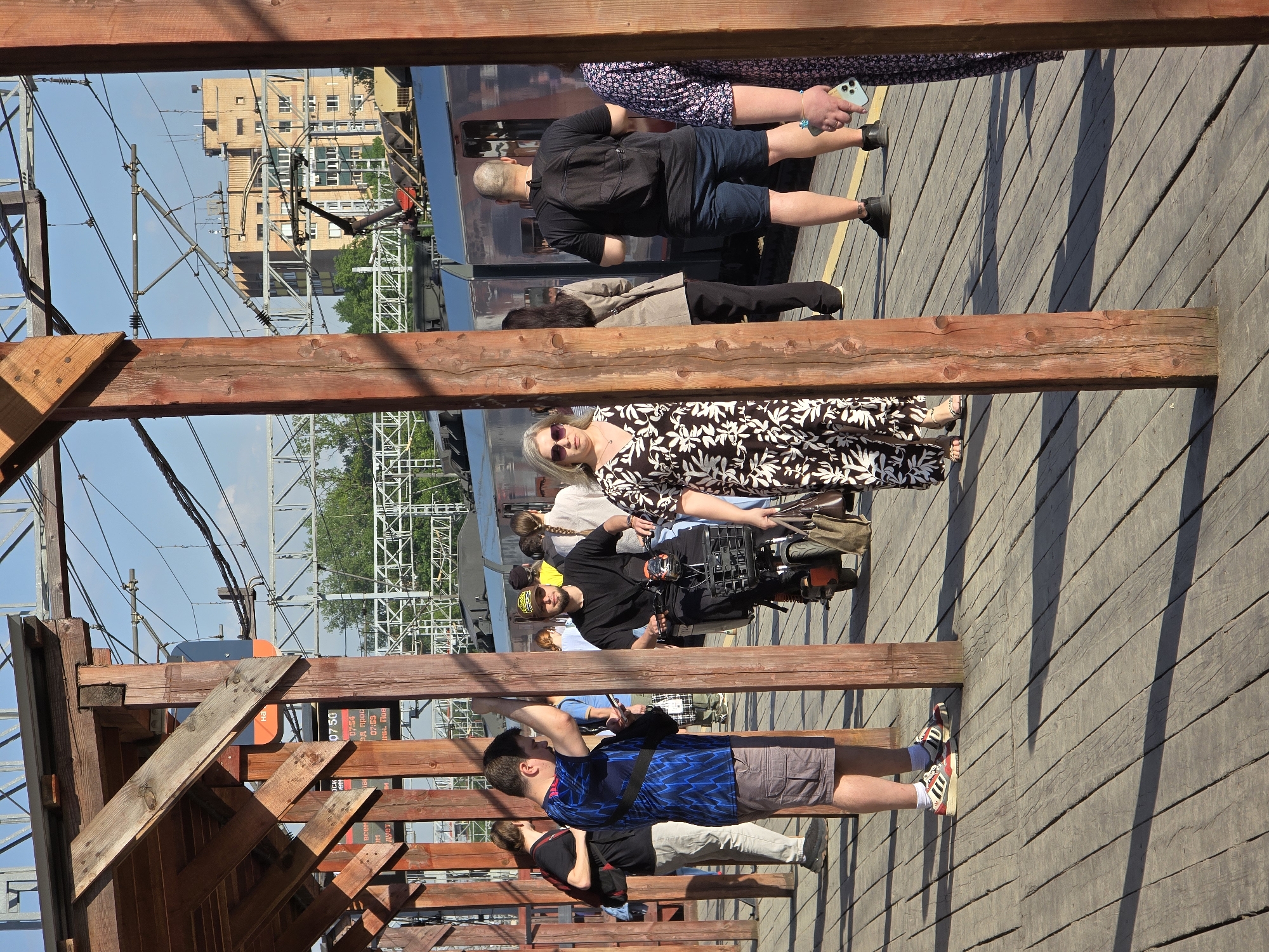 Captured from a low-angle perspective, the image depicts a group of people standing on a wooden platform, potentially a train platform, in Khimki, Russia. The composition is framed by wooden beams and supports, adding a structural element to the scene.
Several individuals are present: one man wearing blue shirt and shorts. One woman wearing a black and white dress and sunglasses, along with others. An individual is operating a film camera.
The backdrop includes a building, and overhead electrical wires and structures. The lighting indicates a sunny day, casting shadows across the wooden platform. The overall impression is that of a public space, possibly a transportation hub, with individuals engaging in various activities.