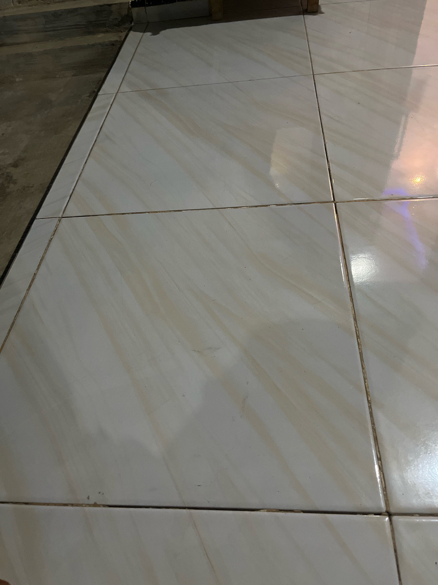The image shows a close-up, high-angle view of tiled flooring. The tiles are rectangular, light-colored, with a subtle marbled pattern of beige and light brown streaks. The grout lines are visible and appear to be slightly discolored, possibly due to age or dirt. To the left of the tiled area, there is a darker, rougher surface that appears to be concrete or dirt. The lighting suggests an indoor environment, with reflections visible on the glossy surface of the tiles. There are no people or specific identifiable objects other than the flooring itself. The scene is devoid of any activity. This image is of a floor in Kebumen, Indonesia.