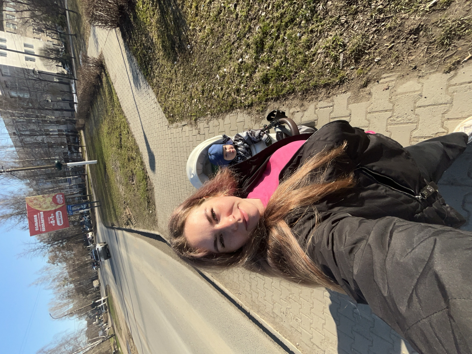 Here is a detailed description of the image:
**People:** The image features a woman taking a selfie with a baby in a stroller.
**Setting:** The location appears to be a sidewalk alongside a street in the city of Chaikovsky, Russia. This is determined by the presence of a billboard for "ESHE PIZZA" which offers free pizza as a part of a "2+1" deal and states "Большая ветчинка и гривы".
**Objects:** A baby stroller, a billboard, a street with cars, buildings, trees, and a sidewalk with paving stones are visible.
**Action:** The woman is taking a selfie with her phone, capturing herself and the baby in the stroller, along with the surrounding urban environment. The baby is seated in the stroller.
**Environment:** The environment is a typical urban scene with buildings, trees, and traffic. The weather is sunny.
**Composition:** The image is taken from a slightly low angle, showing the woman, the baby, and a section of the cityscape. The focus is on the woman and baby, but the background provides context.
