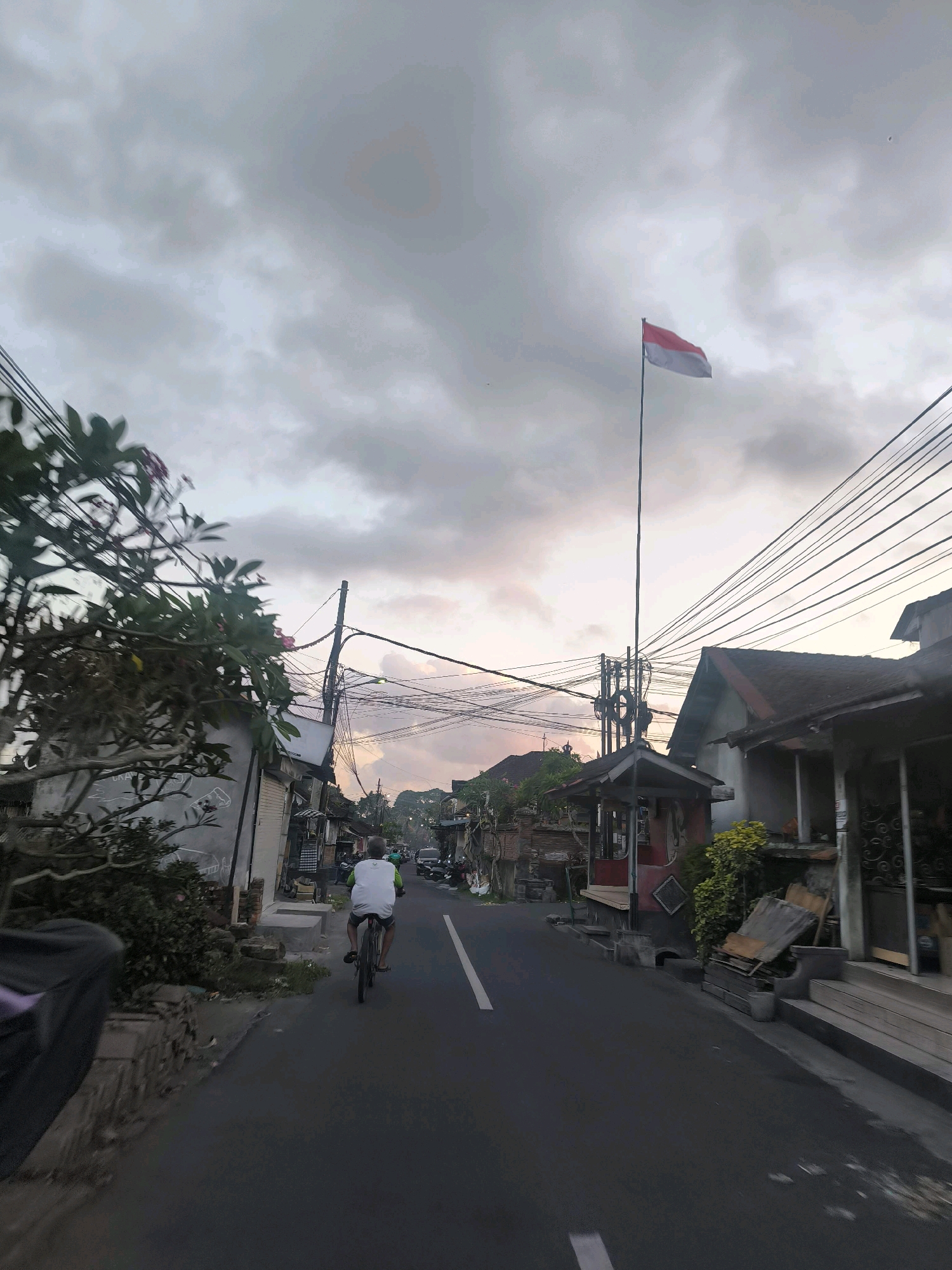 A person rides a bicycle down a paved road in Tabanan, Indonesia. The sky is overcast with dramatic clouds tinged with sunset colors. A flag, red and white, flies from a tall pole on the right side of the road. Buildings line both sides of the street, with power lines crisscrossing overhead. Trees and vegetation are visible on the left side of the road. The scene appears to be a residential or commercial street during twilight.