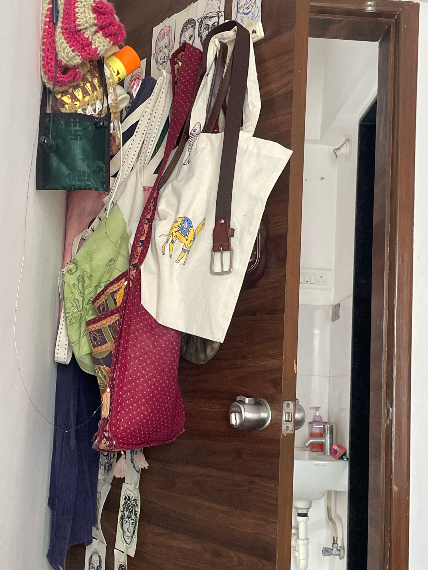 In Loni Kalbhor, India, a wooden door is ajar, revealing a glimpse into a room. On the door's surface and hanging from hooks are an assortment of bags and items. These include a dark green patterned bag, a light green patterned bag, and a deep red bag with a white polka dot pattern. A white tote bag with a brown leather strap and a yellow camel graphic is also visible. Other items include a knitted item in pink and cream, a yellow and orange cylindrical object, and several pieces of paper with hand-drawn faces. The wall to the left of the door is white. The area visible through the open doorway shows a white tiled wall, a white sink with a chrome faucet, a purple pump-action bottle, and a red object on the sink.