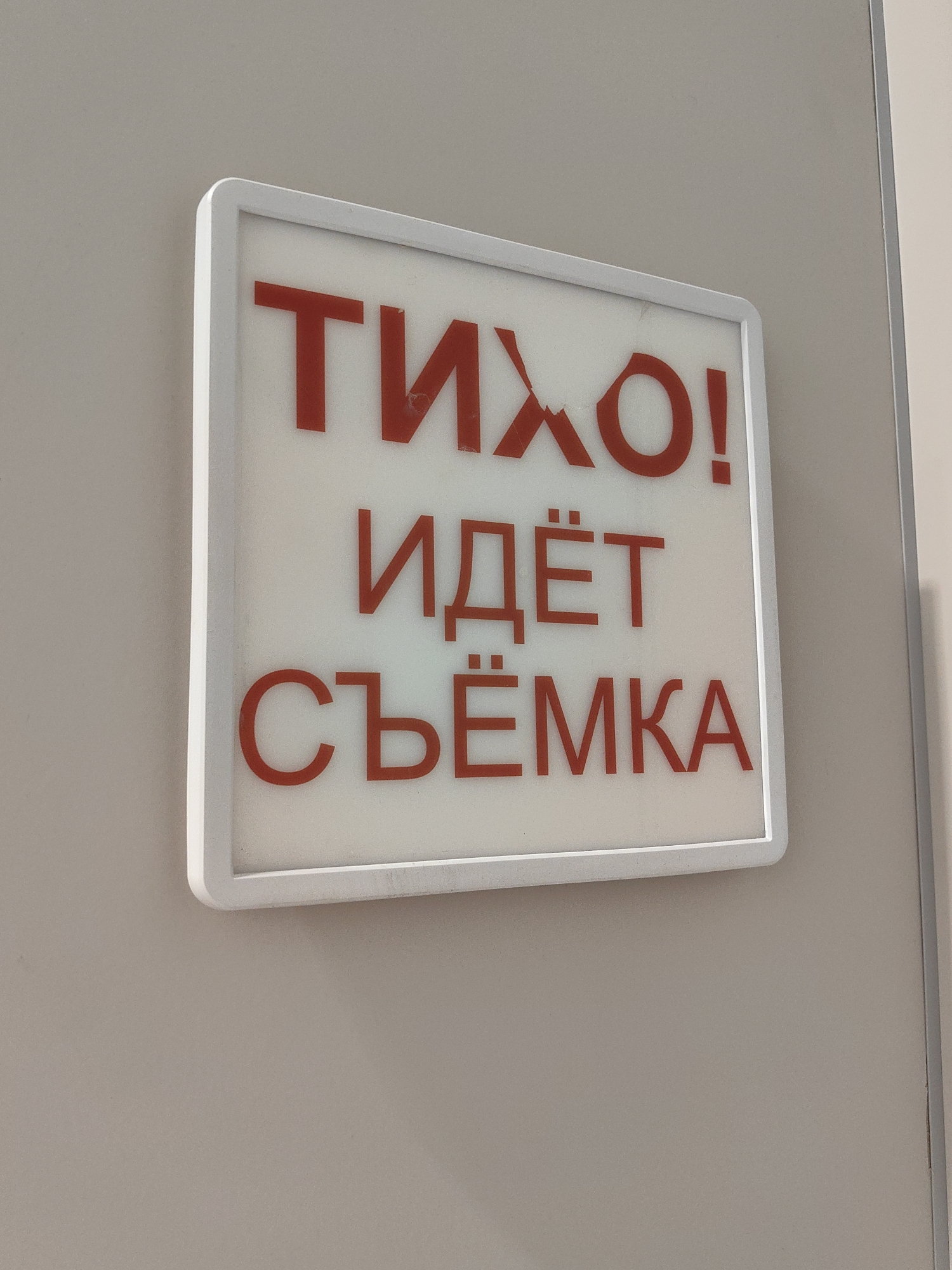 A white illuminated sign with red text is mounted on a light gray wall. The text on the sign reads "ТИХО! ИДЁТ СЪЁМКА". This translates to "QUIET! FILMING IN PROGRESS." The sign is rectangular with rounded corners and a white border. The wall appears to be a smooth, matte surface. The lighting suggests an indoor environment. There are no people or other objects visible in the frame.