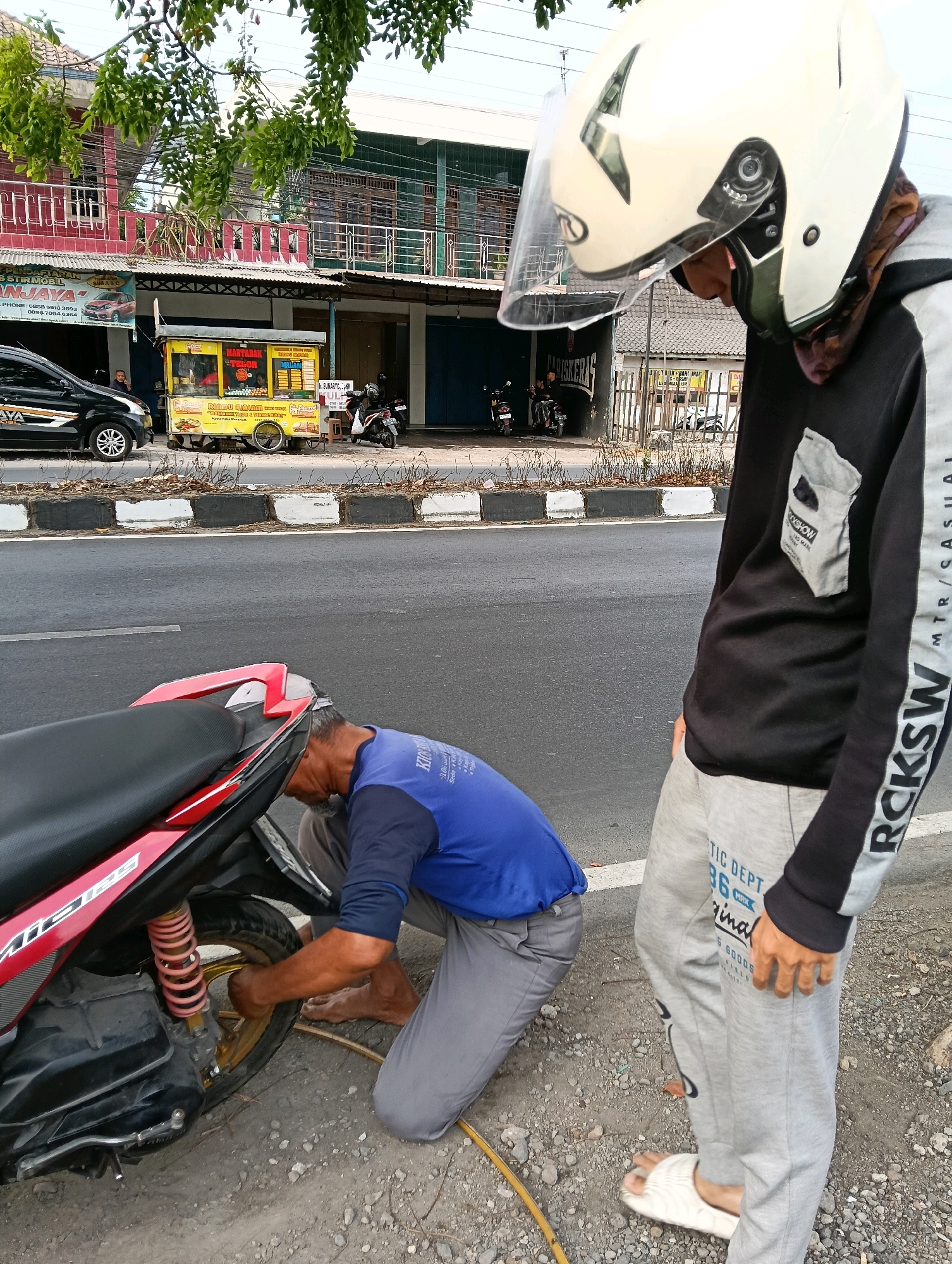 The image depicts an outdoor scene in Jaten, Indonesia, where two men are near a red and black motorcycle on the side of a road. One man, wearing a blue shirt and grey pants, is crouched down and appears to be working on the rear wheel of the motorcycle, possibly inflating the tire with a yellow hose. The other man, standing and wearing a white helmet, black and grey hoodie, and grey pants, is looking down towards the work being done. In the background, there are buildings with storefronts, a yellow food cart, and parked motorcycles and a car. The environment suggests a commercial or roadside area.