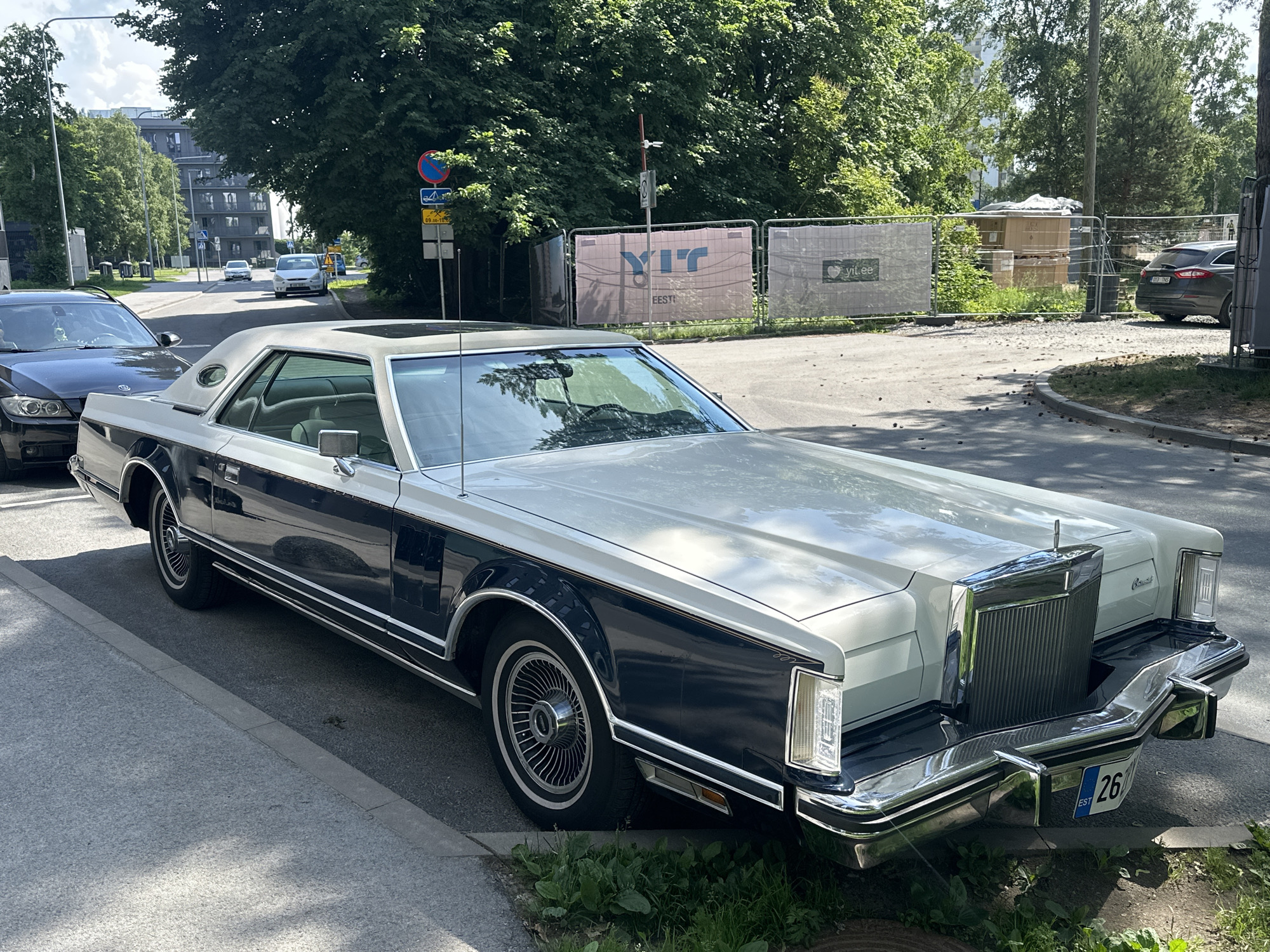 A two-tone Lincoln Continental Mark V, primarily silver and dark blue, is parked on the side of a street in Tallinn, Estonia. The car is viewed from the front-right quarter. The vehicle has a prominent chrome grille and bumper. The license plate is visible and reads "EST 26". Across the street, a construction site is visible with white and pink banners that say "YIT" and "yit.ee". Trees line the street, and a modern apartment building is visible in the background, along with street signs and other parked cars. The day is sunny.