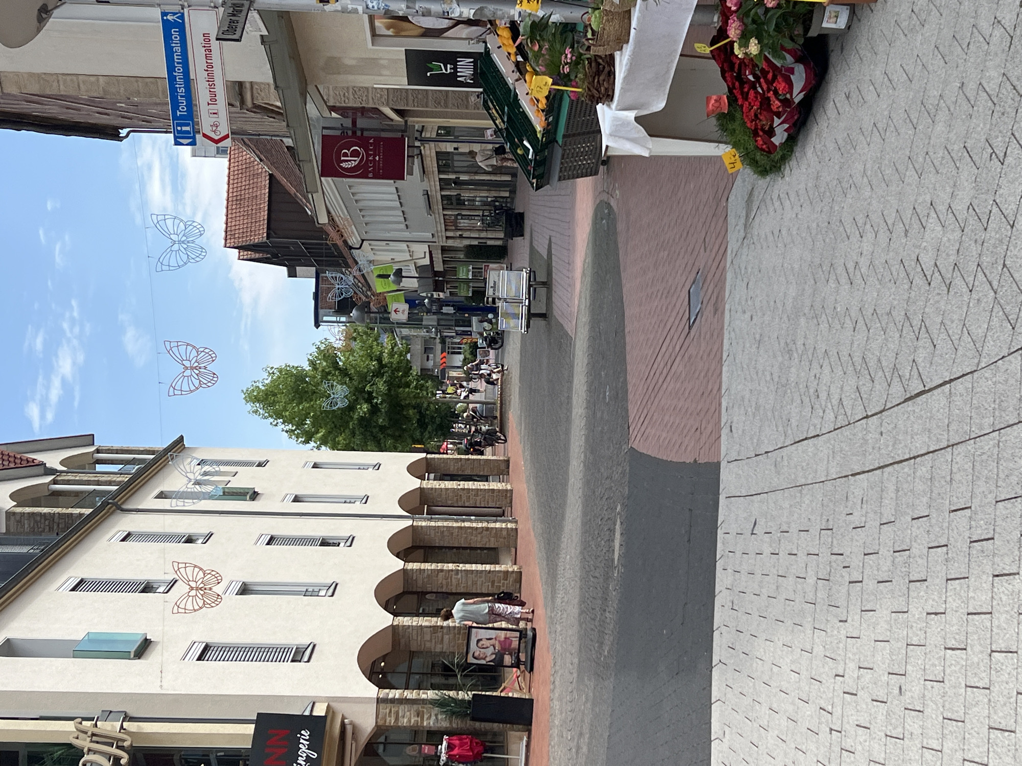 The image captures a street scene in a town with buildings lining both sides of a pedestrian walkway. The architecture suggests a European setting, with the buildings featuring a mix of light-colored facades and arched ground-floor entrances. Decorative butterfly-shaped lights are strung across the street.
On the left, a building with a sign that says "NN lingerie" is visible. A woman in a light-colored top and patterned pants is walking away from the camera, in front of a display featuring images of models. Further down the street on the left, a few people are seen walking, and a motorbike is parked.
On the right side of the street, there are shops with awnings and displays. One sign reads "BACKECK" with a "B" logo. Further down, a stall is set up with a white tablecloth, displaying an assortment of fruits and flowers. A sign with "AMIN" and a shopping cart logo is also visible on a building.
The street is paved with bricks in a reddish-brown hue, transitioning to gray cobblestones in the foreground. The sky is partly cloudy, with a bright blue sky visible. There are trees and streetlights present along the walkway. The overall impression is a lively, outdoor commercial area.