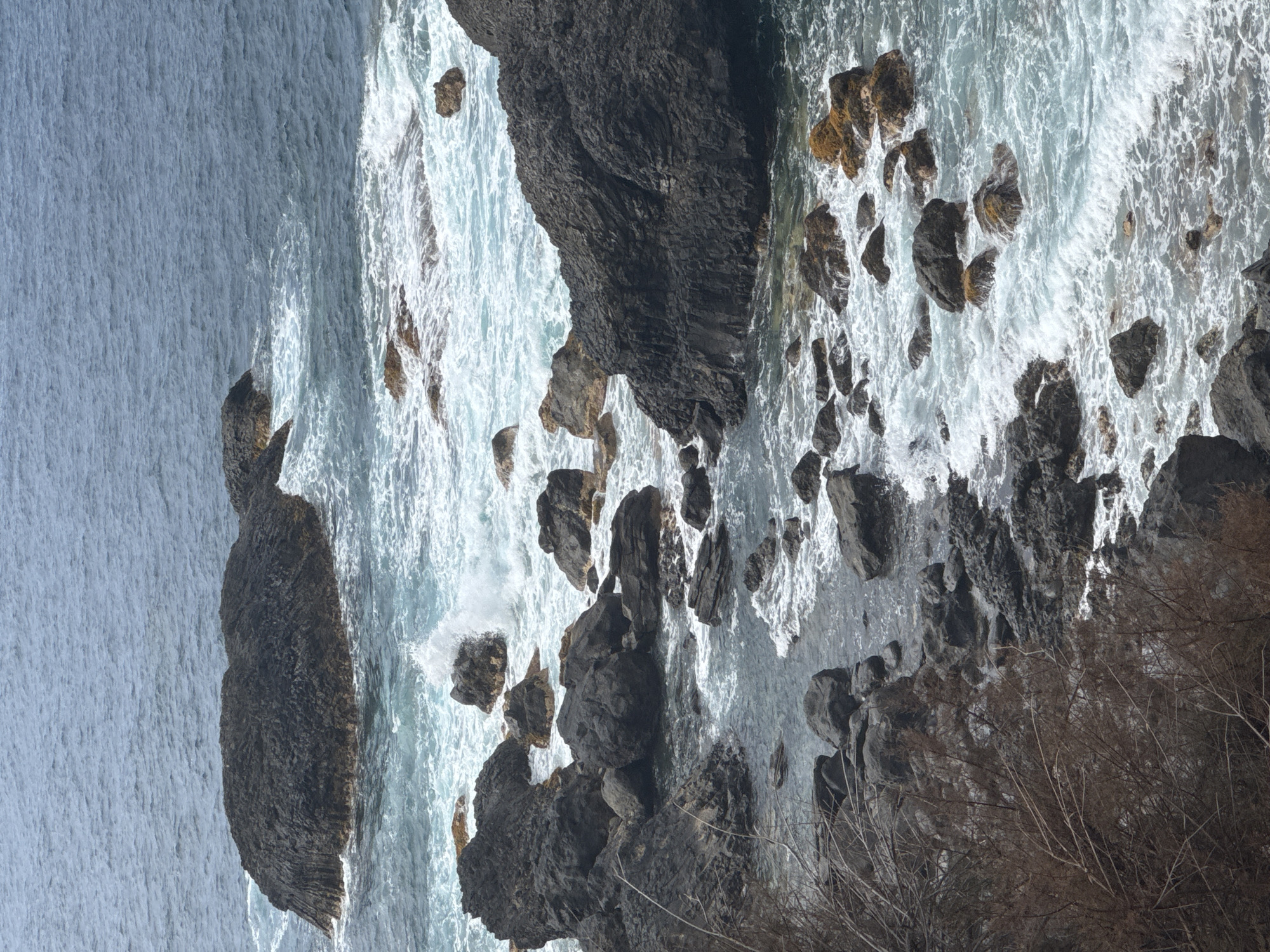 A high-angle, medium shot captures a dynamic seascape with dark, jagged rocks protruding from the turbulent, teal-colored ocean. The waves crash against the rocks, creating white foam and spray that add to the chaotic, yet beautiful, scene. The water's surface is rippled, indicating wind or currents. In the foreground, the rocky coastline is visible, with dry, bare branches of a shrub extending from the left edge of the frame. The overall impression is one of raw, natural power and the relentless force of the sea.