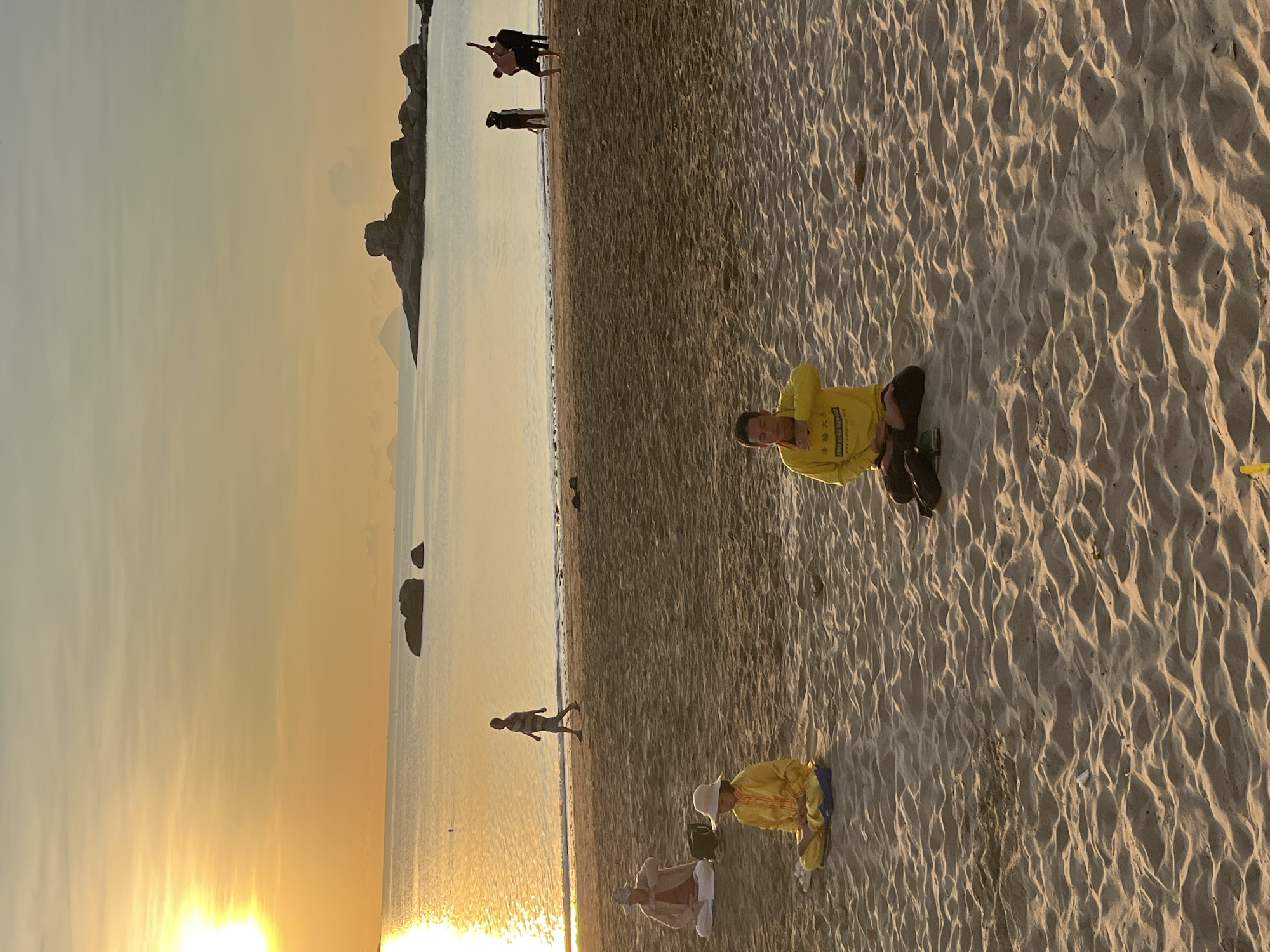 A photograph taken in Bac Nha Trang Ward, Vietnam, shows a sandy beach at dusk. The sky is a hazy yellow. To the left are some rocky formations in the sea. A few figures are visible in the distance, near the water's edge. Closer to the foreground, a person in a yellow top is seated in the sand. Further down the beach, another person is kneeling, also wearing yellow. Other individuals are sparsely scattered along the beach.