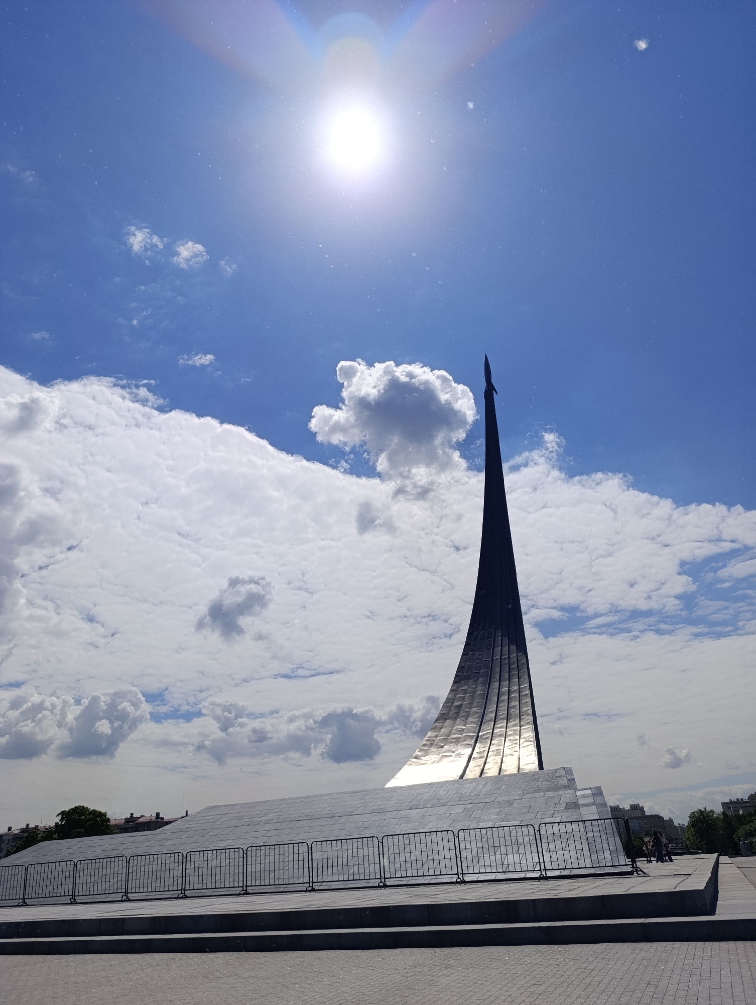 The image displays the iconic Monument to the Conquerors of Space (Cosmonauts Monument) in Moscow, Russia, under a brilliant blue sky. The central subject is the colossal, upward-sweeping obelisk, clad in gleaming titanium panels, which reflects the bright sunlight. A stylized rocket sits atop the monument, pointing skyward. The monument's base is a substantial, angled structure, also in reflective material.
The sky is a vibrant blue, dotted with fluffy white cumulus clouds. The sun is prominently visible in the upper left of the frame, appearing as a bright white disk and creating a noticeable lens flare and light streaks. Numerous tiny specks, likely dust on the camera lens, are scattered across the clear sky, particularly noticeable in the brighter areas.
In the foreground, black metal crowd control barriers are positioned along the base of the monument, separating it from the paved plaza. The ground features light-colored interlocking pavers, indicative of a public square. In the distance, behind the monument's base, faint silhouettes of urban buildings and some green trees are visible, suggesting an urban setting.
A few small figures of people can be seen on the right side, near the base of the monument, appearing to be visitors or passersby observing the structure. The scene captures a clear, sunny daytime, likely late morning or early afternoon, emphasizing the monument's impressive scale and reflective surfaces against the vivid sky. There is no visible text in the image.