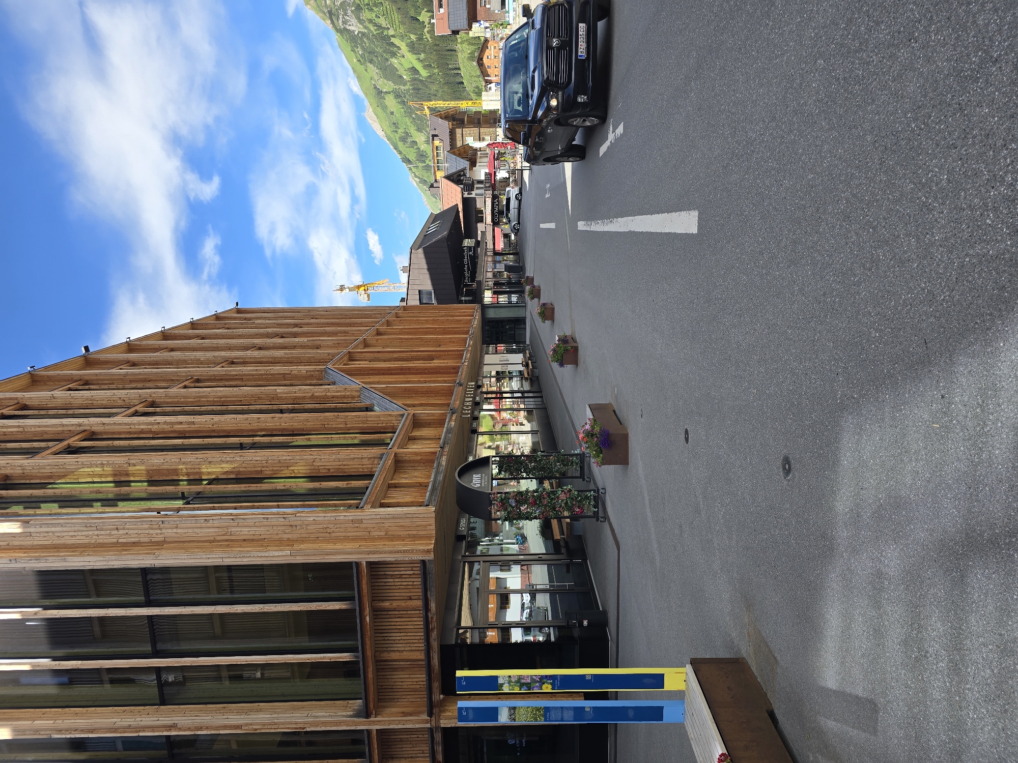 The image shows a street scene in Lech, Austria, on a sunny day. A modern building with a wooden facade lines the left side of the street, featuring large windows and vertical wooden beams. In front of the building, there are shops with glass entrances and awnings. To the left, two tall, narrow display stands, one blue and one yellow, are positioned on the sidewalk. A dark-colored pickup truck is driving on the asphalt road in the right lane, with white lane markings visible. In the background, the town of Lech is nestled in a valley, with traditional Austrian architecture and lush green, tree-covered mountains under a blue sky with scattered white clouds. A yellow construction crane is visible in the distance. The scene appears to be in a pedestrian-friendly area with planters containing flowers.