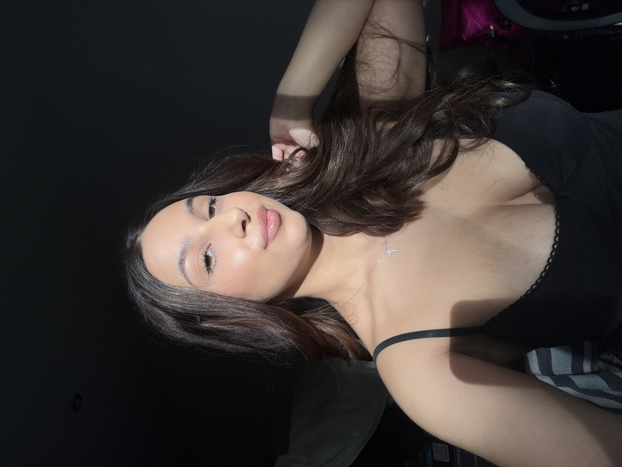 A woman is lying down, posing for a selfie. She has long, dark brown hair and is wearing a black top. A silver necklace with a cross pendant is visible on her neck. The background is dark, with a strip of light across her face. A ring light and other miscellaneous items are partially visible in the background. The location is Mönchengladbach, Germany is not discernible from the image.