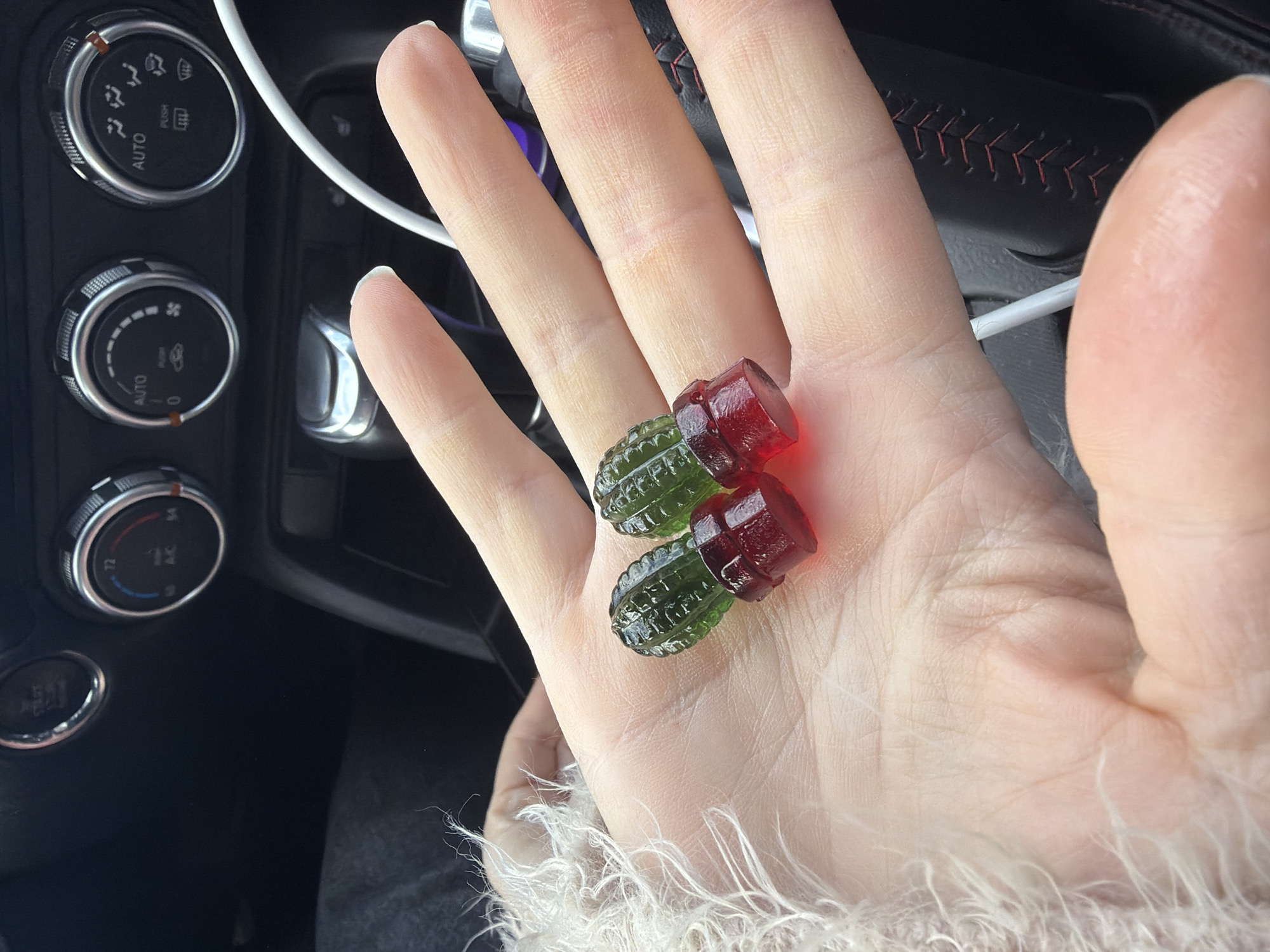 The image displays a close-up of a person's open hand holding two gummy candies, set against the interior of a car. The candies are distinctly shaped like small grenades, one translucent green and the other translucent red. The hand is light-skinned, showing visible creases and skin texture. Around the person's wrist or lower arm, a fluffy, light-colored material, resembling faux fur, is visible.
In the background, part of a car's dashboard is visible, featuring two circular climate control knobs with metallic trim. These knobs include options such as "AUTO," symbols for air distribution (face, feet, defroster), and fan speed. One knob displays the number "72," likely indicating a temperature setting. A white cable, possibly a charging or auxiliary cord, cuts across the upper portion of the frame. To the right, a dark surface with red stitching, possibly part of a steering wheel or gear shifter, is partially visible. The lighting suggests it is daytime. While the car interior details are generic, the provided context indicates the photograph was taken in Saint Petersburg, Russia. The scene captures a casual moment, possibly on the go.