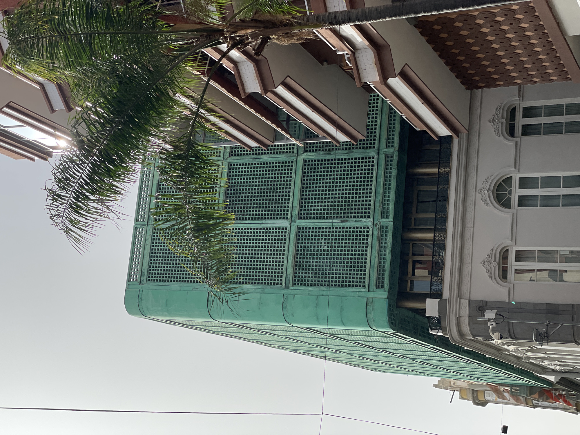 A low-angle shot features a light green building with a squared grid pattern facade in Santa Cruz de Tenerife, Spain, adjacent to a white building with ornate window details. A palm tree stretches across the upper left quadrant, its leaves partially obscuring the structures. Visible architectural features include protruding window sills and an intricate brown tile pattern. Surveillance cameras and electrical wiring are attached to the buildings. The sky is washed out, typical of a bright day. Lower buildings are seen at the bottom right of the frame.
