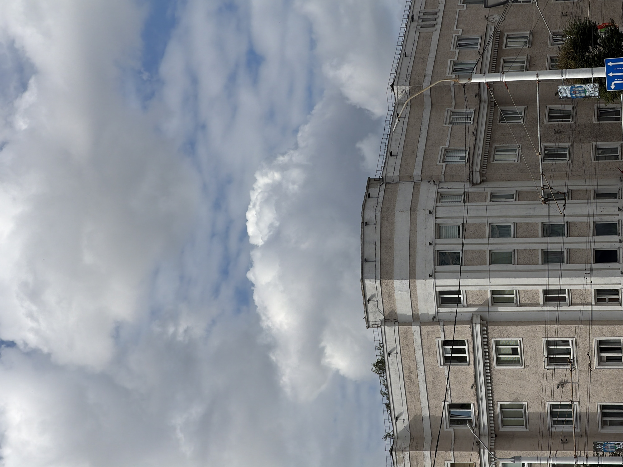 The image shows a multi-story building with a light-colored facade and horizontal beige stripes, located in the city of Mahilyow, Belarus. The building has numerous windows, some with white frames and others with blinds. A corner of the building is visible, featuring vertical white decorative elements. Overhead power lines crisscross in front of the building. In the foreground, a traffic sign with blue arrows indicating directions is present. The sky above is filled with large, dramatic white clouds against a blue background. The scene appears to be a street view in an urban environment.