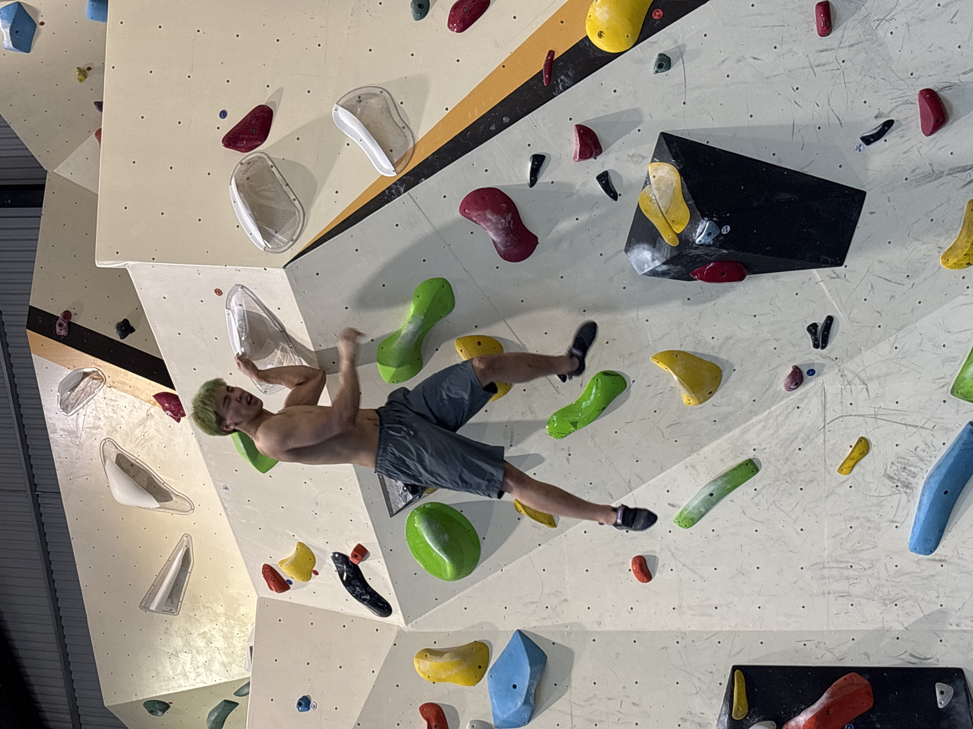 The image shows a young man with dyed green hair bouldering indoors. He is shirtless, wearing dark grey shorts, and climbing on a textured wall with numerous colorful holds of various shapes and sizes. The wall is part of an indoor climbing gym, characterized by its geometric design and artificial climbing structures. The man is mid-climb, with his left arm extended upwards and his right arm bent, appearing to be in the process of reaching for a hold. His facial expression suggests exertion. The gym environment is visible, with the ceiling structure and additional climbing walls in the background. The lighting is artificial, typical of an indoor sports facility. The image does not provide any specific visual cues to indicate its location within Đà Nẵng, Vietnam.