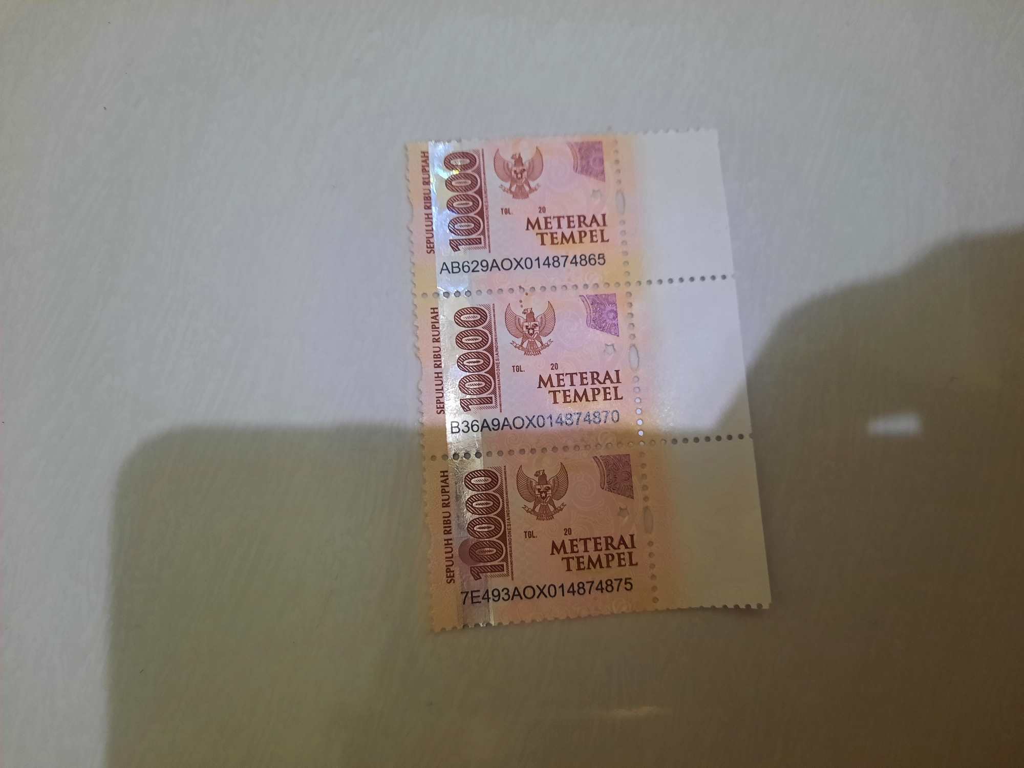 The image shows three postage stamps with the value "10000" and the text "METERAI TEMPEL" printed on them. Each stamp also features an image of Garuda Pancasila, the national emblem of Indonesia, and a unique alphanumeric code. The stamps are arranged vertically, with a perforating line separating each one. The background is a light-colored surface, possibly a table, with a shadow cast on the right side. The image does not provide any information about the location in Lhokseumawe, Indonesia.