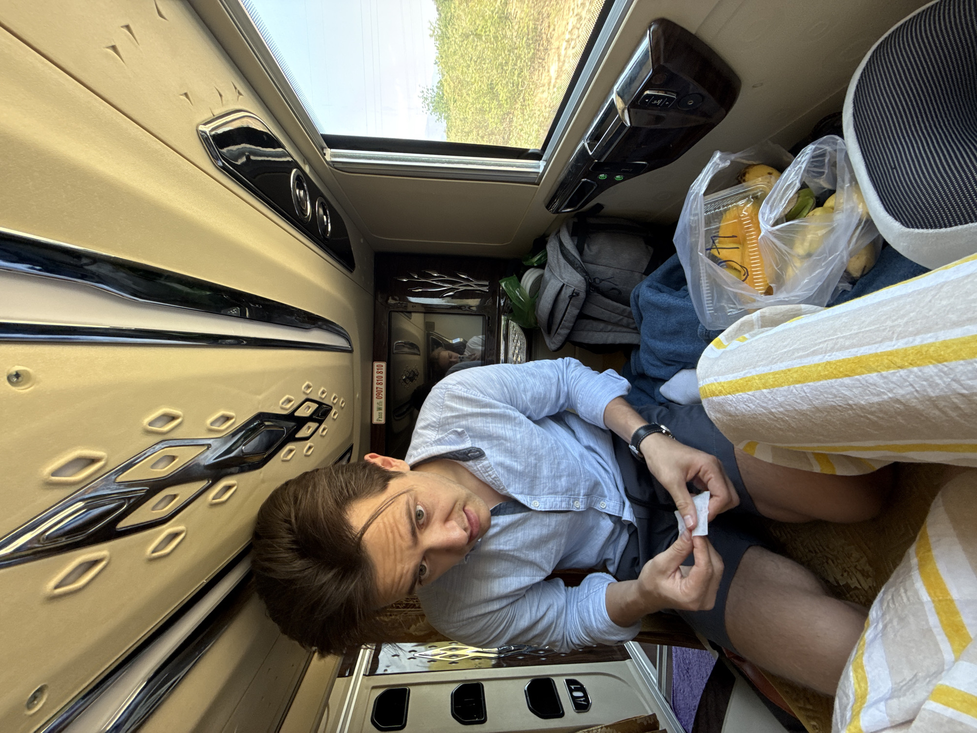 The image depicts a young man inside a compact, individual compartment of what appears to be a sleeper bus or customized travel vehicle, likely in Phuoc Huu, Vietnam. The scene is illuminated by bright natural daylight streaming through an overhead window or sunroof, revealing green foliage and a clear sky outside, indicating it is daytime.
The subject is a young man with short dark brown hair and light eyes, positioned at a slightly oblique angle, looking directly at the camera with a neutral, slightly curious expression. He is dressed in a light blue long-sleeved button-up shirt and dark shorts, wearing a black watch on his left wrist. He is sitting or reclining in his bunk, holding a small, crumpled piece of white paper or tissue in his hands.
The compartment's interior features light beige upholstered walls with decorative chrome accents and raised patterns. Above the man, an overhead panel with controls (possibly for lights or air conditioning) is visible. Adjacent to him, on bedding with yellow and white stripes, are a clear plastic bag holding bananas and other fruit in a container, a grey backpack, and some blue clothing. A striped headrest or cushion is also present. A small screen or mirror below the man displays his reflection.
On a panel near the man's head, there is text written in Vietnamese: "PASS: Win 0907 113 810," which appears to be a phone number. The overall impression is of a traveler resting comfortably in a personalized space during a journey.