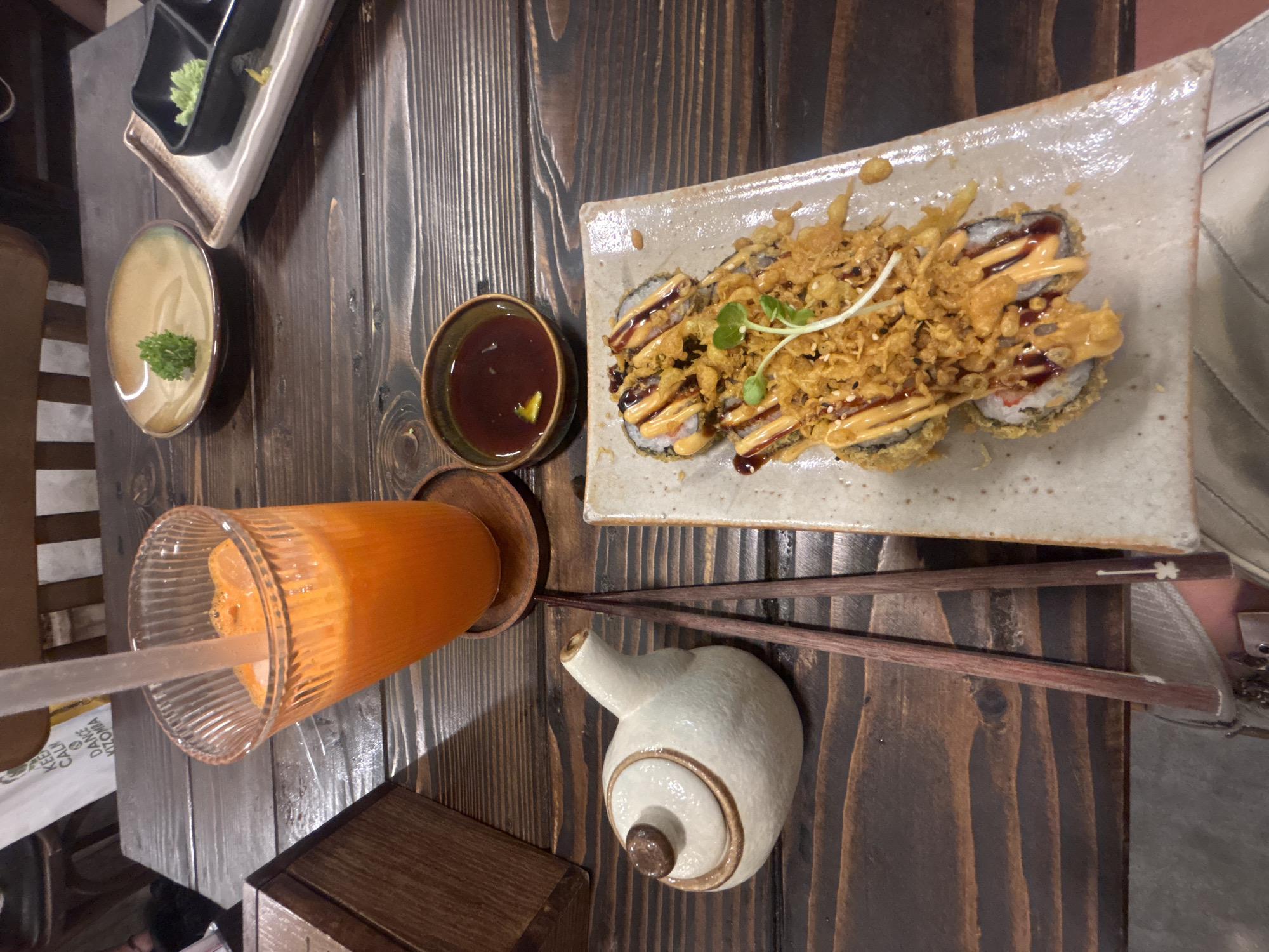 A close-up, eye-level shot captures a table setting at a restaurant in Da Nang, Vietnam, featuring a meal and beverage. In the foreground, a light-colored ceramic teapot, resembling a snail with a brown knob and rim, sits to the left. Next to it are a pair of dark red chopsticks with white tips. To the right, a rectangular ceramic plate holds two pieces of deep-fried sushi rolls, generously topped with crispy fried onions and a drizzle of spicy mayonnaise and dark sauce, garnished with a small green sprout.
In the mid-ground, a tall, ridged glass filled with vibrant orange juice and ice sits on a wooden coaster. A clear plastic straw extends from the glass. Adjacent to the juice is a small, dark brown ceramic bowl containing a dark sauce. Further back, two small plates are visible on the wooden table; one is a light brown, shallow bowl with a small sprig of green garnish, and the other is a divided black and white dish, with one compartment holding green wasabi paste.
The table itself is made of dark, weathered wood planks with a visible grain. In the upper left corner, a portion of a wooden box is visible, and behind it, a white paper bag with text, including the words "KEEP CALM," "DANCE KIZOMBA," and "ĐÀ NẴNG," suggests the location. The background shows parts of chairs and possibly other tables, indicating an indoor dining environment. No people are clearly visible in the frame, but a person's arm wearing a watch is partially visible at the bottom edge of the image, suggesting someone is seated at the table. The lighting appears to be ambient and slightly dim.