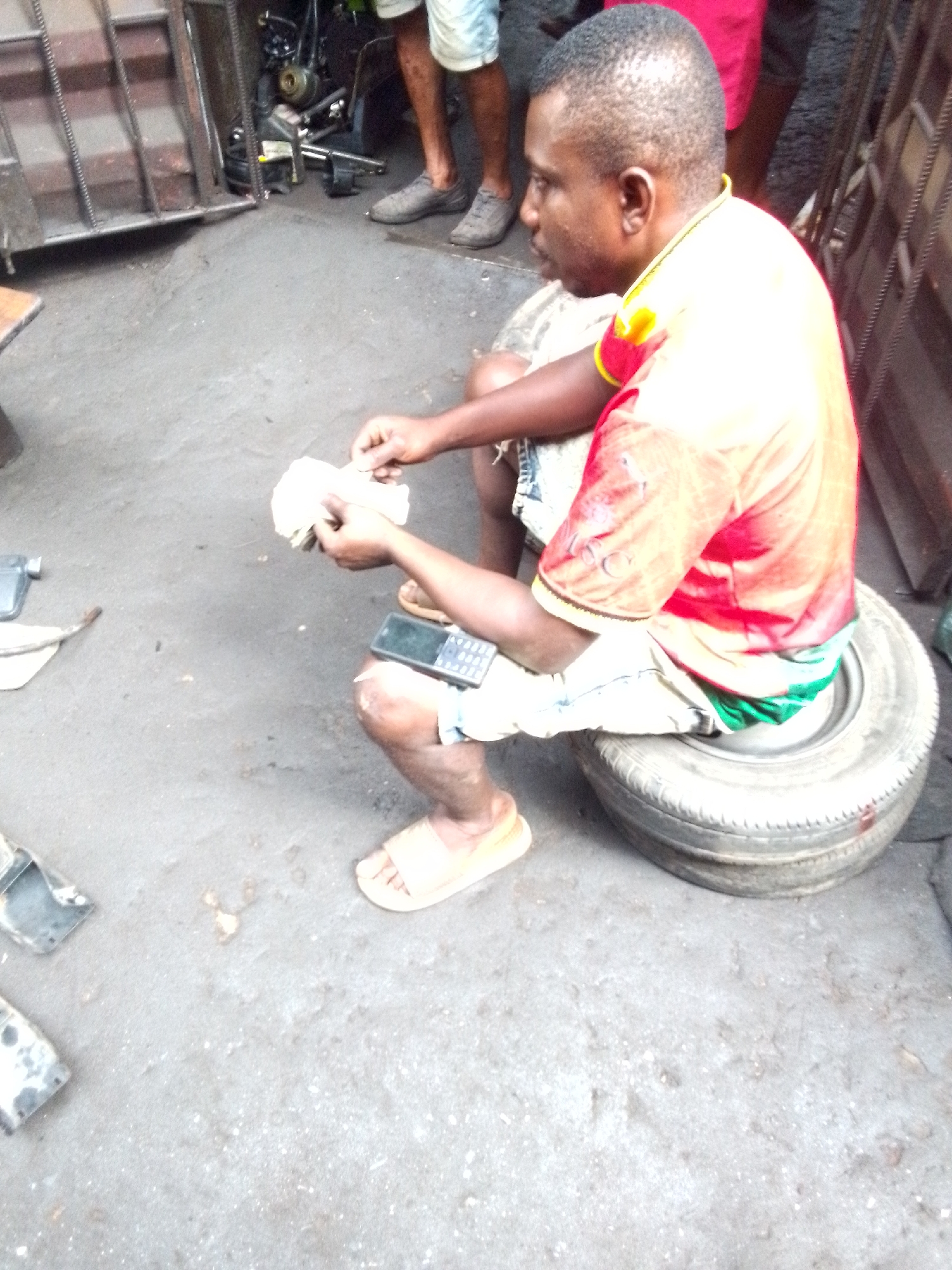 An overhead-angle shot captures a man sitting informally outdoors in what appears to be an urban or semi-urban setting in Onitsha, Nigeria. The man, with dark skin and short, closely-cropped hair, is positioned on a worn, flat car tire serving as a makeshift seat. He is wearing a red and yellow short-sleeved t-shirt, light-colored shorts, and light brown slip-on sandals.
His focus is directed downwards towards his hands, where he is holding and seemingly counting or organizing a wad of papers or currency notes. A black feature phone with a physical keypad rests on his left thigh. The ground beneath him is a rough, grey surface, possibly concrete or packed dirt, with scattered debris.
In the background, to the left, a dark metal gate or grill structure is visible, suggesting an entrance to a building or enclosure. Further back, obscured by the gate, are parts of what looks like a motorcycle or engine. To the right and upper background, legs and feet of another person, wearing grey shoes, are partially visible, along with a glimpse of red fabric. The overall scene suggests a casual, everyday activity in a busy, functional area, likely during the daytime. No distinct text is visible in the image.