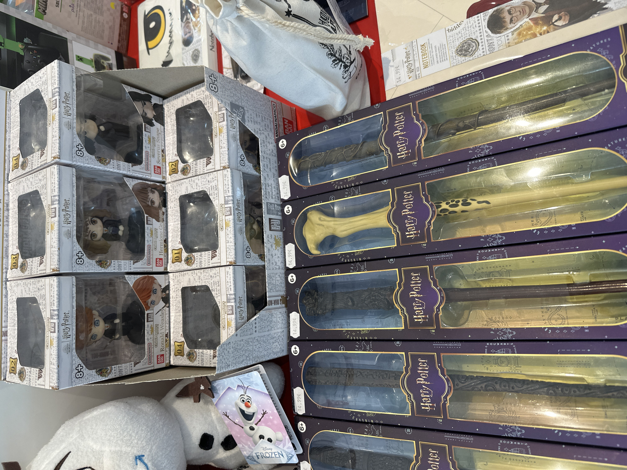 The image displays a retail display of Harry Potter merchandise in what appears to be a store in Calais, France. The primary focus is on two rows of boxed Harry Potter wands, with some chibimaster figures in the background. The wands are in dark purple boxes with gold accents, each featuring a clear plastic window showcasing the individual wand. The figures are in smaller, white boxes, also with clear windows, depicting characters like Ronald Weasley and Severus Snape. To the left, a plush toy, possibly Olaf from Frozen, is visible. In the background, other merchandise such as notebooks and a drawstring bag can be seen. The overall environment suggests a retail setting, likely a toy or souvenir shop.