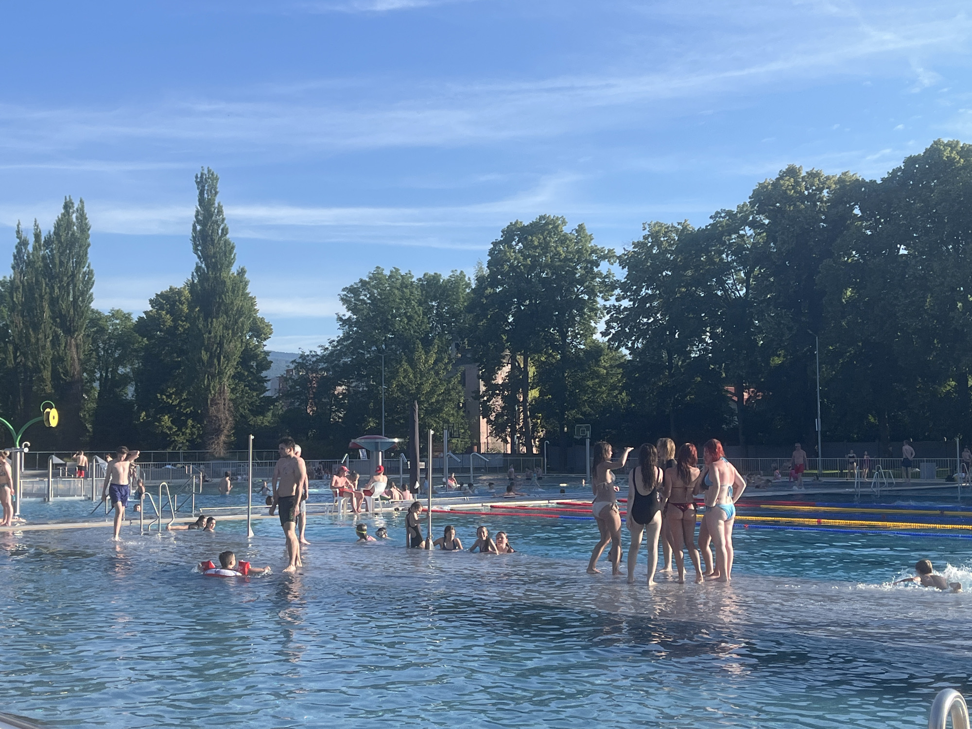 The image depicts a busy public swimming pool on a clear, sunny day. In the foreground, several people are enjoying the water, including a child with a floatation device and a group of women in swimsuits standing on a raised platform. The middle ground features more swimmers and a young boy in the water. In the background, there are trees, buildings, and a blue sky with wispy clouds. There are also lane markers in the pool and lifeguard chairs visible. The overall scene suggests a recreational setting with people engaged in various water activities.