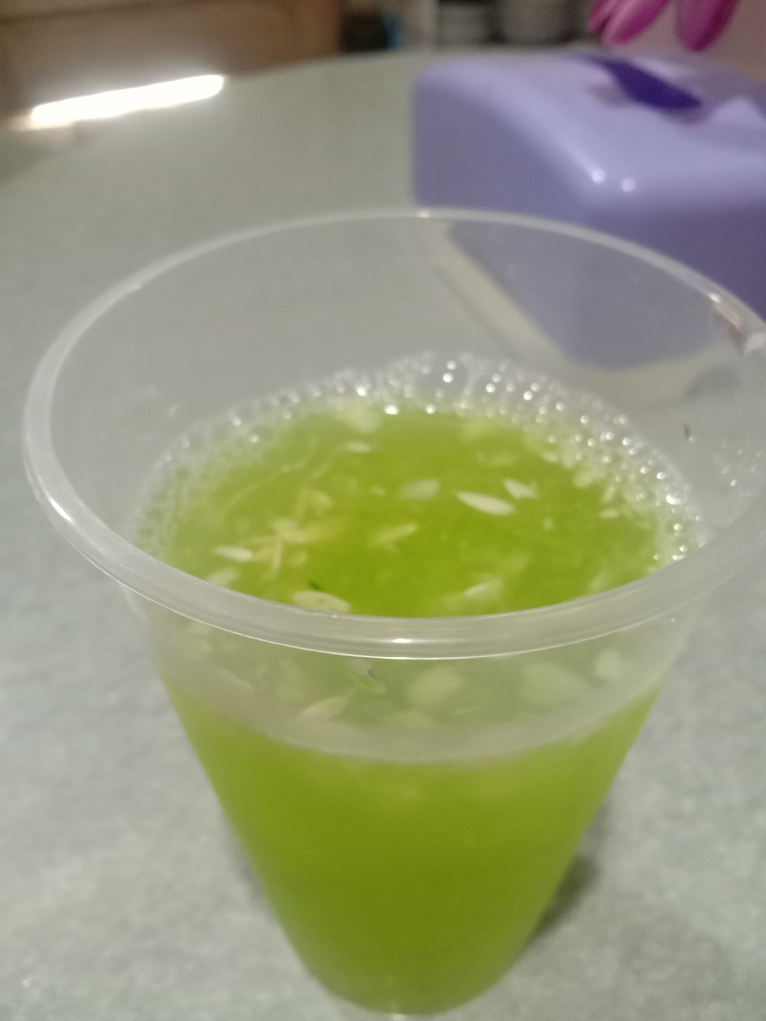 A close-up shot shows a clear plastic cup filled with a green liquid, likely a drink, with small, light-colored solids floating within it. The drink appears to have a frothy or bubbly surface. The cup sits on a light-colored surface, possibly a table or countertop. In the background, out of focus, is a light purple rectangular object. The image does not provide information about the location in Lhokjok, Indonesia, or any individuals present.