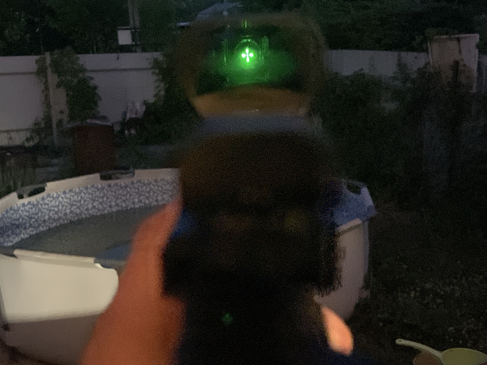 The image, taken at night in Vasilyevo, Russia, shows a first-person perspective through a firearm's optical sight. The sight displays a green reticle with a central dot and four radiating lines. In the foreground, a hand with an orange hue, likely from artificial lighting, is visible holding the firearm. The background is dark and shows a blurry swimming pool with a blue and white patterned liner, and a white fence. To the right of the pool, there is dense foliage and some indistinct structures.