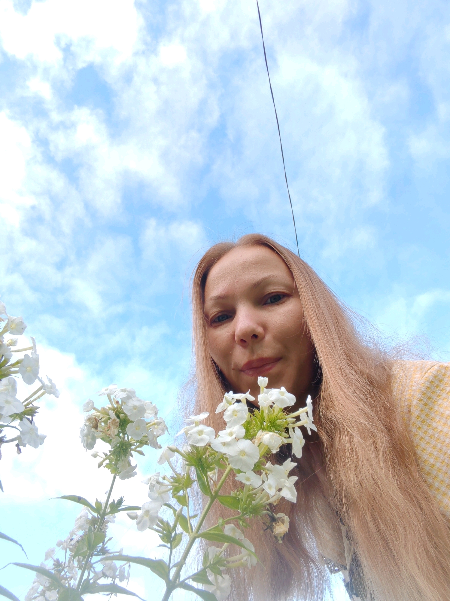 The image shows a low-angle, close-up view of a woman with long, light brown hair and fair skin, looking towards the camera. She is wearing a yellow and white checkered top. In the foreground, there are several clusters of small white flowers on green stems. The background consists of a bright blue sky with scattered white clouds, and a thin black wire is visible diagonally across the upper portion of the frame. The image appears to have been taken outdoors in a garden or natural setting, possibly in Чухлэм, Russia, given the context provided.