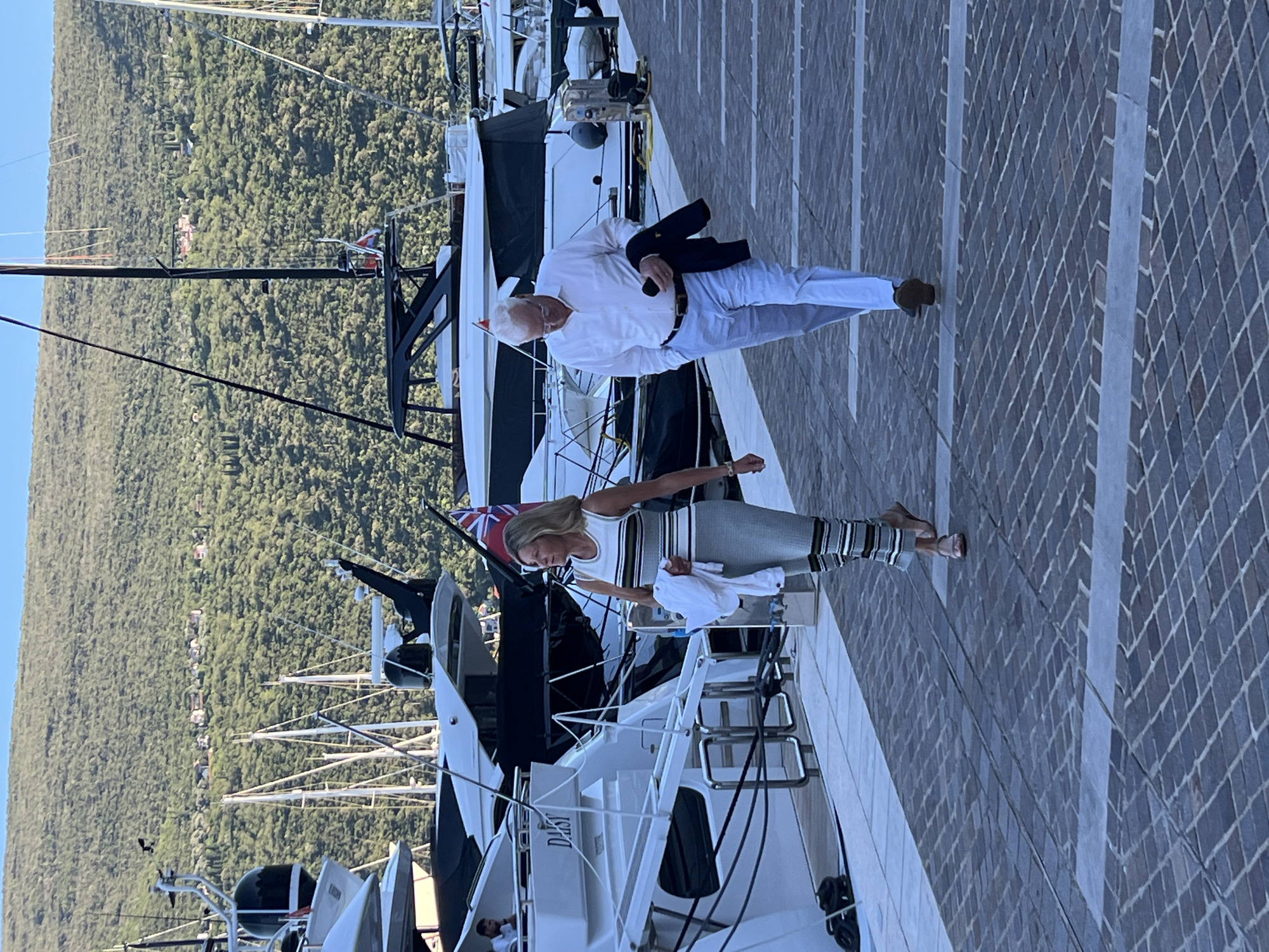 This image shows a man and a woman walking on a stone-paved promenade in the city of Herceg Novi, Montenegro. They are walking alongside a marina filled with yachts. The woman, on the left, is wearing a striped sleeveless dress and holding a white jacket. The man, on the right, is wearing a white dress shirt, light-colored pants, and holding a dark jacket. In the background, a lush green hillside dotted with buildings rises above the marina, with a clear blue sky overhead. The overall scene suggests a warm day in a coastal resort town.