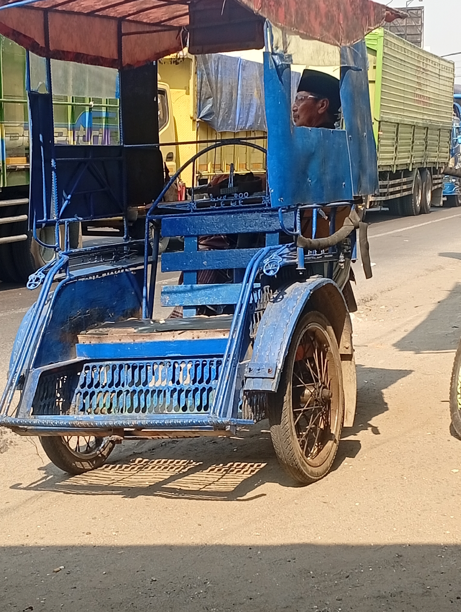 In Bajingjowo, Indonesia, a blue, motorized becak, a three-wheeled passenger vehicle, is driven by a man wearing a black peci hat and glasses. The becak has a retractable canopy made of red fabric and a blue metal frame. It is on a paved road, with the sandy shoulder visible in the foreground. Behind the becak, a large yellow truck and a green and white truck are partially visible, suggesting a roadside environment. The scene is illuminated by bright sunlight, casting distinct shadows on the ground. The becak has a passenger compartment with a wooden seat and a decorative metal grille. The rear wheel of the becak is visible, with its spokes and tire.