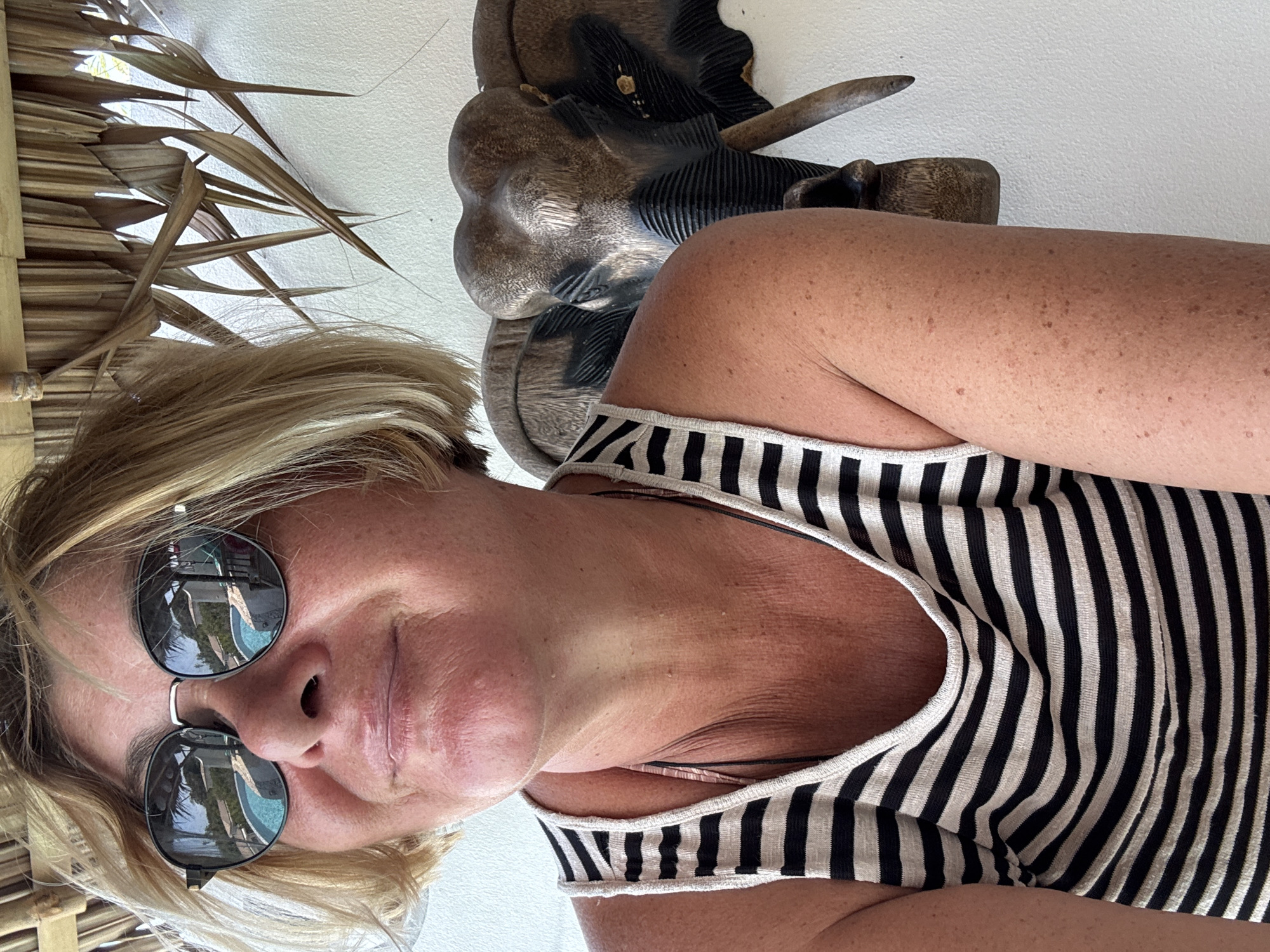 A woman with short blonde hair and sunglasses is positioned in the foreground, wearing a black and white striped tank top. Behind her, there is a decorative wooden carving, possibly depicting an elephant, mounted on a white wall. To the left of the frame, a thatched roof or covering is visible, suggesting an outdoor or semi-outdoor setting. The sunglasses reflect palm trees, a building, and a blue-and-green-patterned body of water. Based on the provided location, the setting is likely a resort or a similar establishment in Baan Chaweng Noi, Thailand.