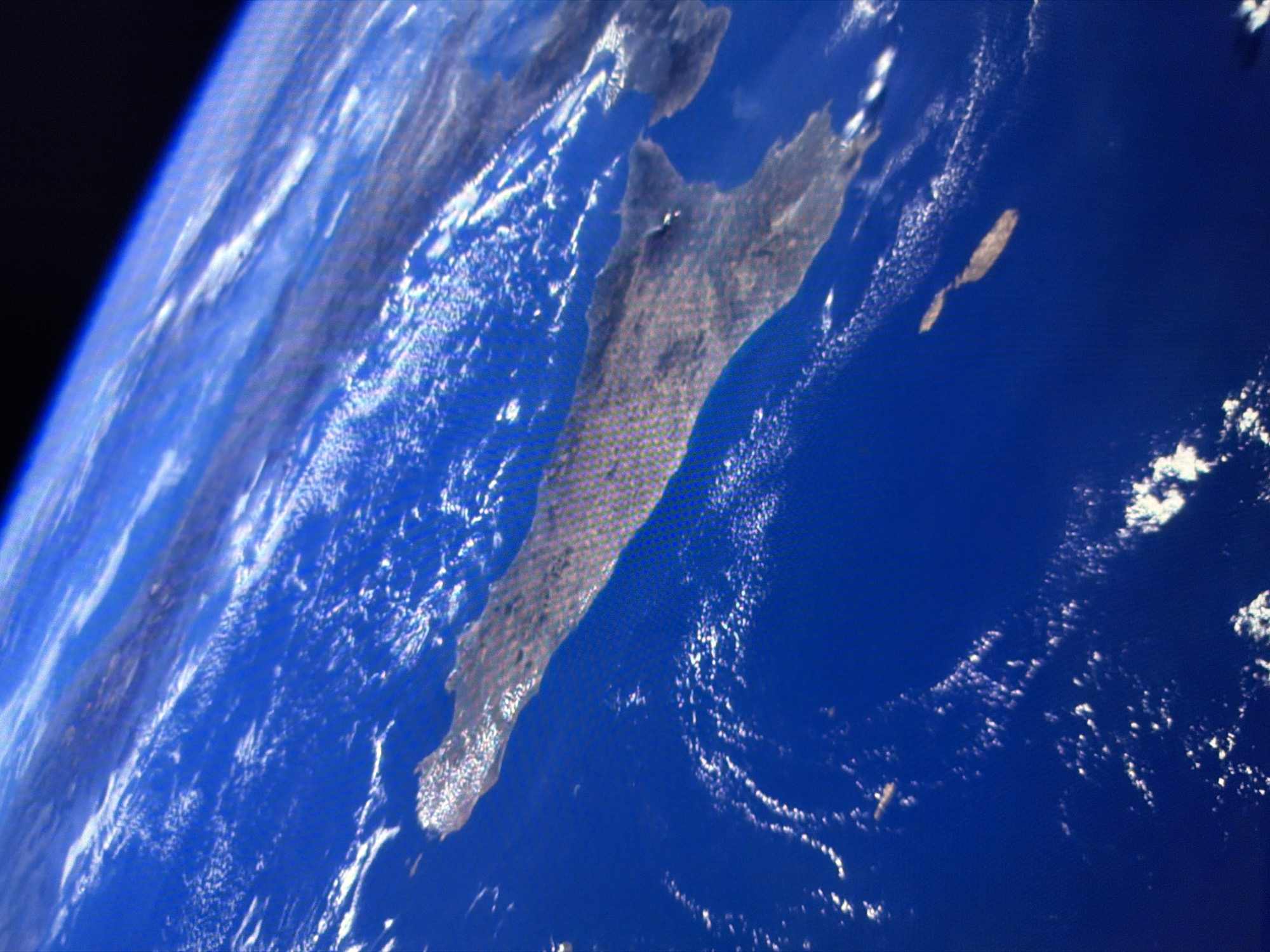 The image shows a view from space, looking down at a body of land surrounded by blue ocean. The landmass is long and narrow, with a textured surface that suggests it is covered in vegetation or buildings. There are also scattered clouds visible on the surface of the water and the land. The image is taken at an angle, giving a sense of perspective and depth. There are no people, subjects, objects, or actions visible in the image. The scene is a geographical landscape from a high vantage point. The location in the city of Kyiv, Ukraine is not identifiable from this image.