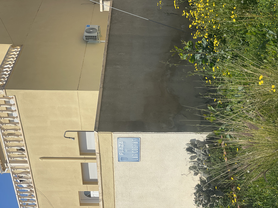 An outdoor, sunlit scene depicts a pale yellow, two-story building in Urbanización Ciudad Quesada, Spain, specifically at "Plaza de las Costas" as indicated by a light blue street sign affixed to a lower cream-colored wall. The building features a white balustrade along its upper roofline. On its lower visible facade, there is a white arched door and two rectangular windows with closed white roller shutters. An external, dark gray shower head fixture protrudes from the wall above the lower cream-colored section. Further right on the building's facade, a white outdoor air conditioning condenser unit with a visible fan is mounted.
A lower, textured concrete wall, primarily dark gray and partially stained, forms a corner in the midground, separating the building from the foreground vegetation. In the foreground, dense green grasses, tall weeds, and numerous small yellow wildflowers dominate the lower portion of the image. Behind the lower cream wall to the left, a cluster of prickly pear cacti (Opuntia species) is visible. The scene is illuminated by bright sunlight, casting distinct shadows on the lower walls and vegetation. No individuals are present, nor are any active interactions or movements depicted.