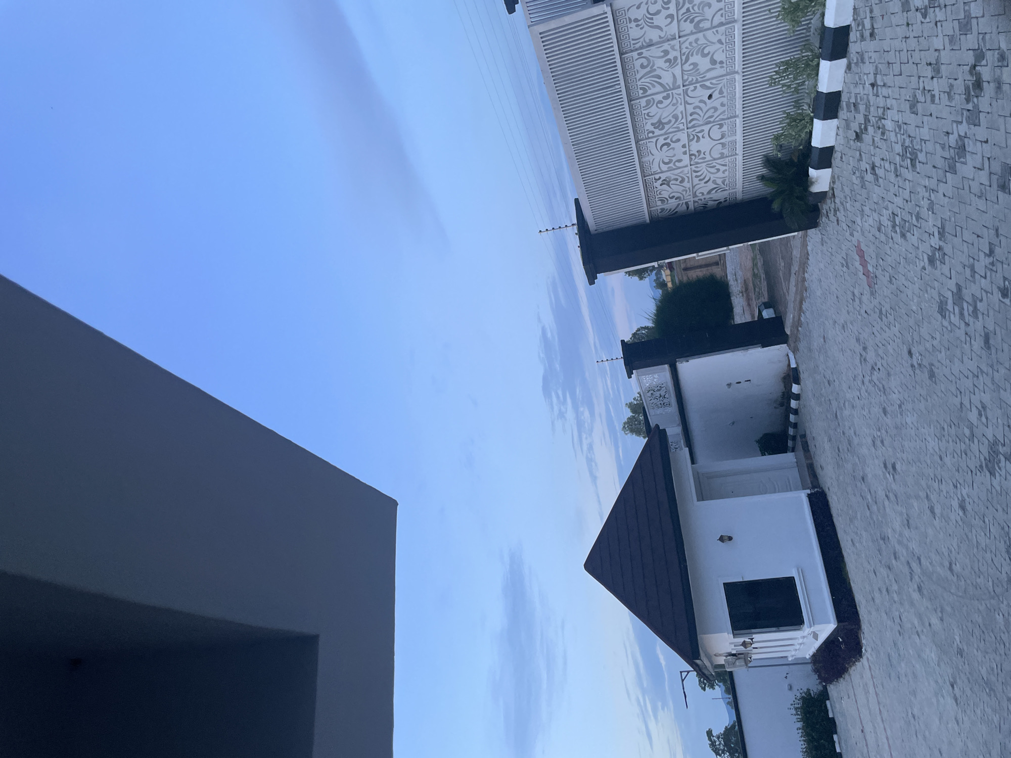 The image captures a street-level view of an entrance to a property in Jauro Yinu, Nigeria. The foreground is dominated by a grey, patterned cobblestone driveway. To the left, a small, white guardhouse with a dark, pyramidal roof is visible. It features a single window with dark glazing and two outdoor lights. A small patch of dark greenery is situated at the base of the guardhouse.
The middle ground is characterized by a white wall and a decorative, tall gate. The gate is predominantly white with intricate black and white patterned panels and black pillars. Security wires are visible atop the gate. Adjacent to the gate, to the left, is an opening leading further into the property, partially obscured by greenery.
The background consists of a clear blue sky with scattered, light clouds. Some trees and foliage are visible beyond the gate and walls, suggesting a suburban or semi-rural setting. The overall impression is of a well-maintained, secured entrance.
