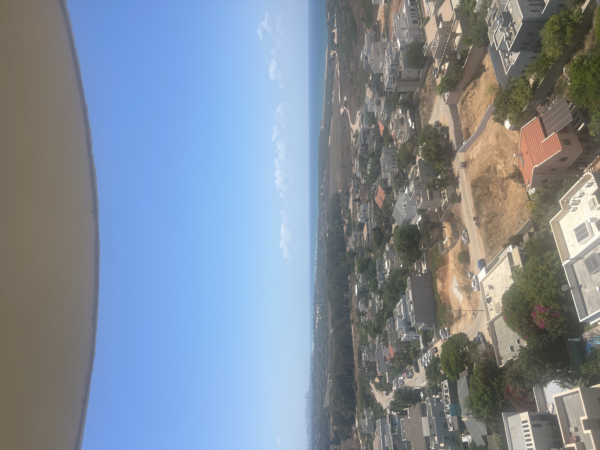 The image presents an aerial view of a residential area in Hadera, Israel, with a backdrop of the Mediterranean Sea and a distant city skyline. The foreground is dominated by the curved edge of what appears to be a balcony or overhanging structure, partially obscuring the bright blue sky. Below this, a dense collection of modern residential buildings with flat roofs and white or light-colored facades stretches across the landscape. The architecture is characterized by clean lines and minimalist design, typical of contemporary suburban development.
Interspersed with the buildings are patches of lush green trees and vegetation, adding a natural element to the urban setting. Roads wind through the neighborhood, lined with parked cars, suggesting an active community. Further in the midground, the terrain becomes more natural, featuring undeveloped land with dirt paths and scrubby vegetation, transitioning towards the coast. The sea is visible as a calm expanse of blue water, meeting a sandy shoreline. In the far distance, a hazy city skyline marks the horizon, indicating the presence of a larger urban center beyond Hadera. The overall impression is one of a well-established, modern residential area situated in a coastal environment.