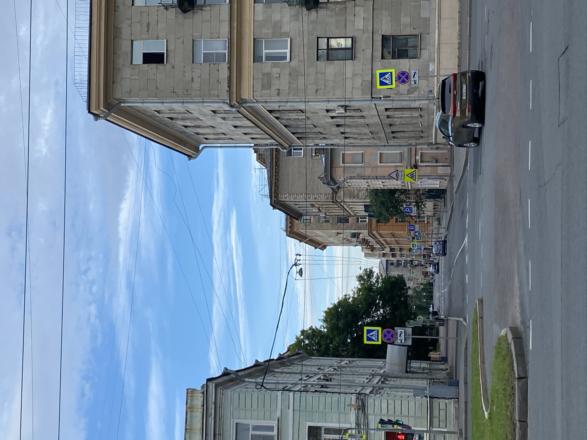 The image shows a street scene in Saint Petersburg, Russia. The perspective is from the street level, looking down a moderately wide street lined with multi-story buildings on both sides. The architecture suggests an older European city. On the right side of the street, a modern, dark olive-colored SUV is turning. The buildings are a mix of styles, with some appearing to be residential or office buildings, characterized by stone facades and numerous windows. The street is paved, with clear lane markings and a pedestrian crossing visible. Several traffic signs are present, including pedestrian crossing signs (blue square with a white pedestrian) and a "no parking" sign. Overhead, power lines crisscross the sky, a common sight in urban environments. The sky is partly cloudy with a mix of blue sky and white clouds. Trees are visible in the background, suggesting the presence of parks or tree-lined streets. The overall impression is that of a typical day in an urban setting.