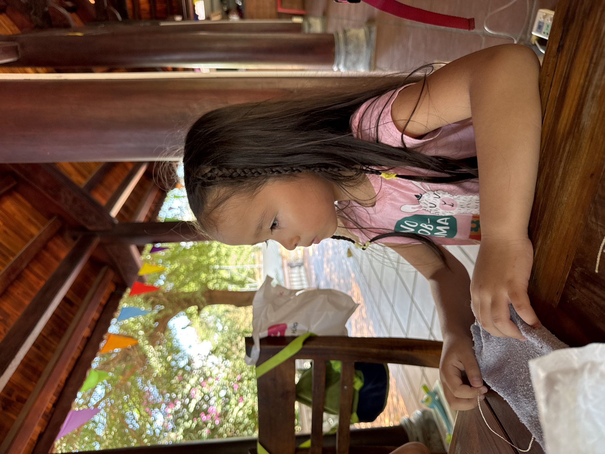 A young girl sits at a wooden table in a structure with a wooden roof, likely a gazebo or covered area. The girl wears a pink shirt with a llama design and has long dark hair with braids. She is focused on something in front of her, possibly crafting or working with materials. Her hands are near a gray cloth or towel and a thin, shiny cord. The outdoor scenery is visible through the structure's opening. There are colorful triangular flags strung up, suggesting a festive or decorative setting. Outside, there is a view of trees and foliage. A chair is visible with a green strap on its back. The location is identified as Mũi Né, Vietnam.