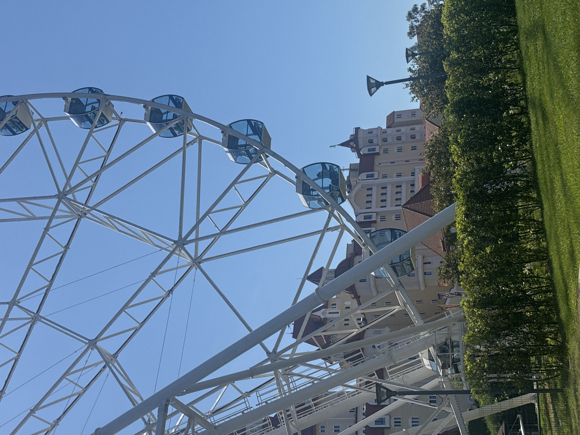 The image displays a large white Ferris wheel prominent against a clear, bright blue sky. The Ferris wheel's structure consists of a complex network of white metal beams and spokes, with several blue-tinted passenger cabins (gondolas) visible along its circumference.
Behind and beneath the Ferris wheel, a multi-story building complex is partially visible. This building features a light-colored facade, possibly beige or cream, complemented by terracotta-colored roofs and architectural accents. It exhibits a distinct style with numerous windows, some arched and some rectangular, along with decorative elements that suggest a resort or themed architectural design, including what appears to be a turret-like structure.
To the right of the building, a dense line of green trees separates the structures from a neatly manicured, vibrant green lawn that fills the right side of the frame. Two dark-colored streetlights, characterized by their unique upward-pointing, triangular light fixtures, are visible; one stands prominently behind the trees, and another is partially obscured by the Ferris wheel and building.
The scene is captured in bright daylight under a cloudless sky, indicating sunny and clear weather. No people are visible, and no specific activity is taking place. The overall setting, featuring a large amusement ride alongside resort-style buildings and well-maintained landscaping, strongly suggests a leisure or entertainment area, consistent with its location in Sirius, Russia. There is no visible text within the image.