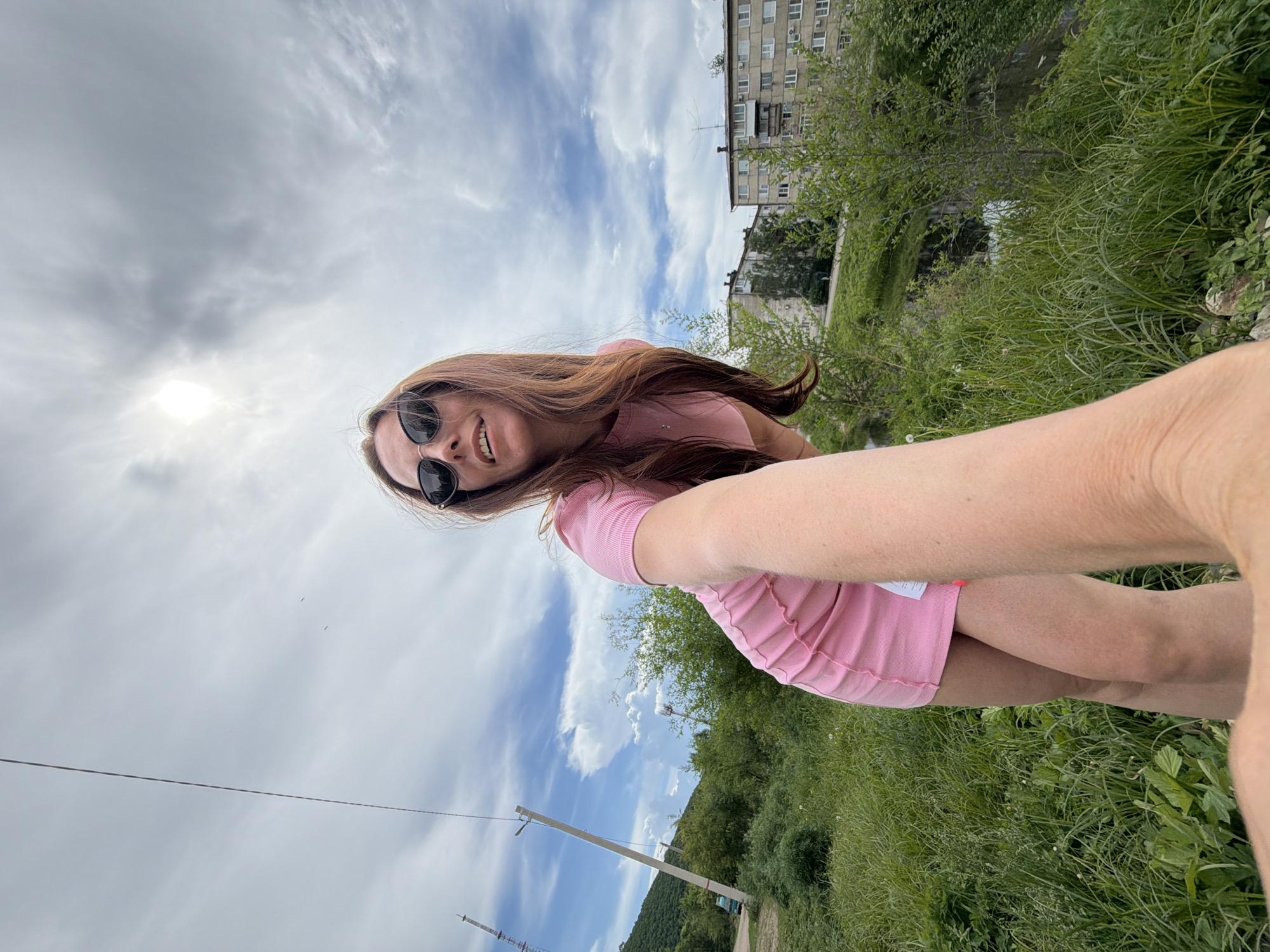 A long-haired woman in a pink dress and sunglasses is taking a selfie outdoors on a cloudy day. She is smiling at the camera, with her arm extended. In the background, there are green trees, tall buildings, and a blue sky with white clouds. The scene appears to be in a suburban area of Birobidzhan, Russia, with a power line and a tower visible to the left.